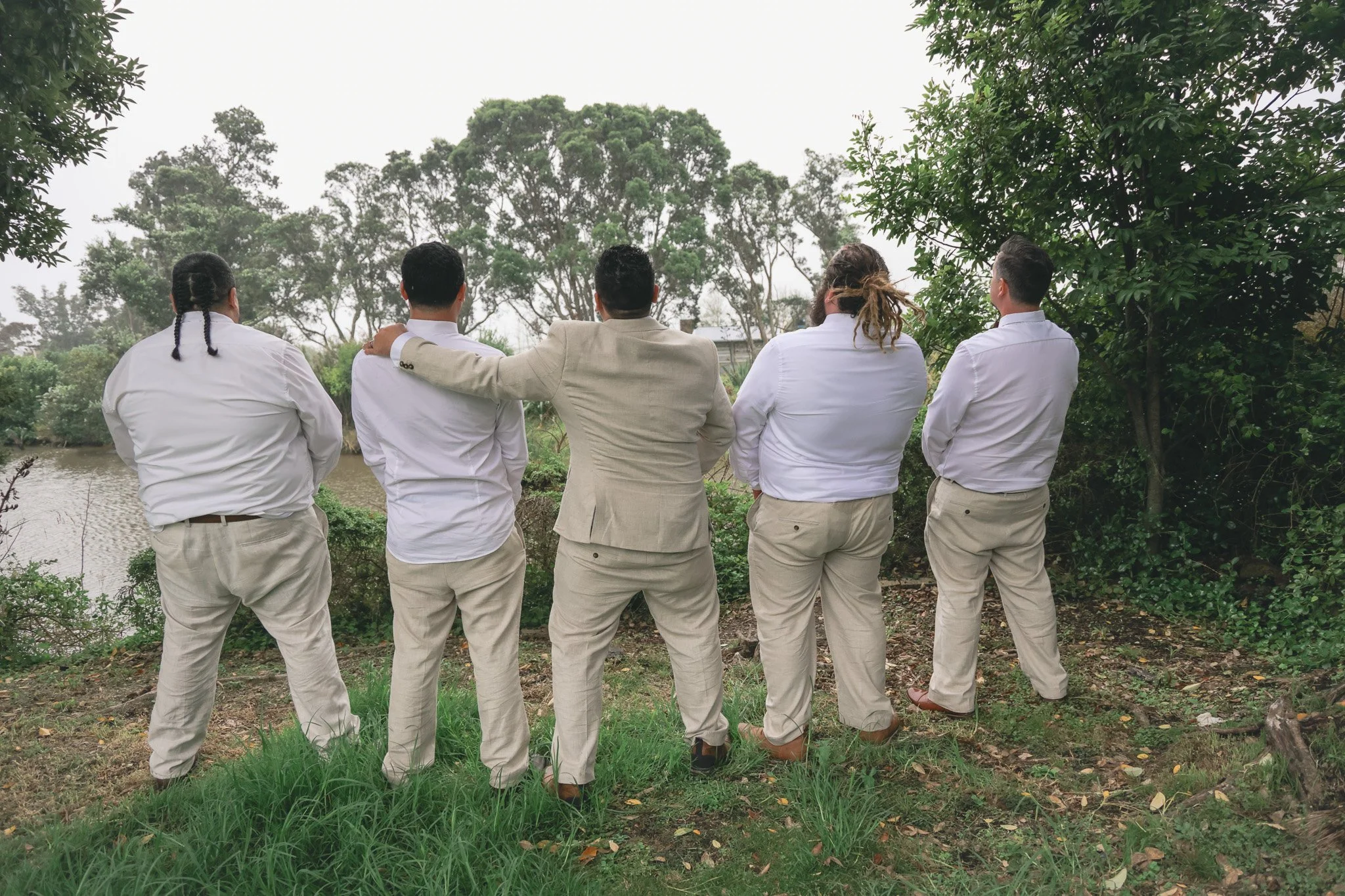 Six people standing outdoors near a water body, facing away, dressed in white and beige clothing, with trees and greenery in the background.