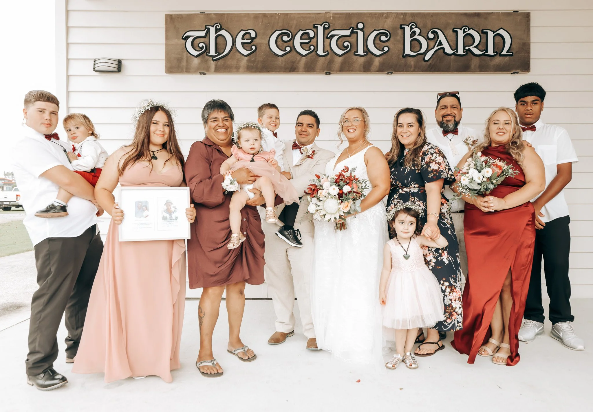 Group of people standing in front of a sign that reads 'The Celtic Barn,' smiling at the camera, dressed in formal and semi-formal attire, holding a bouquet of flowers and a framed photo.