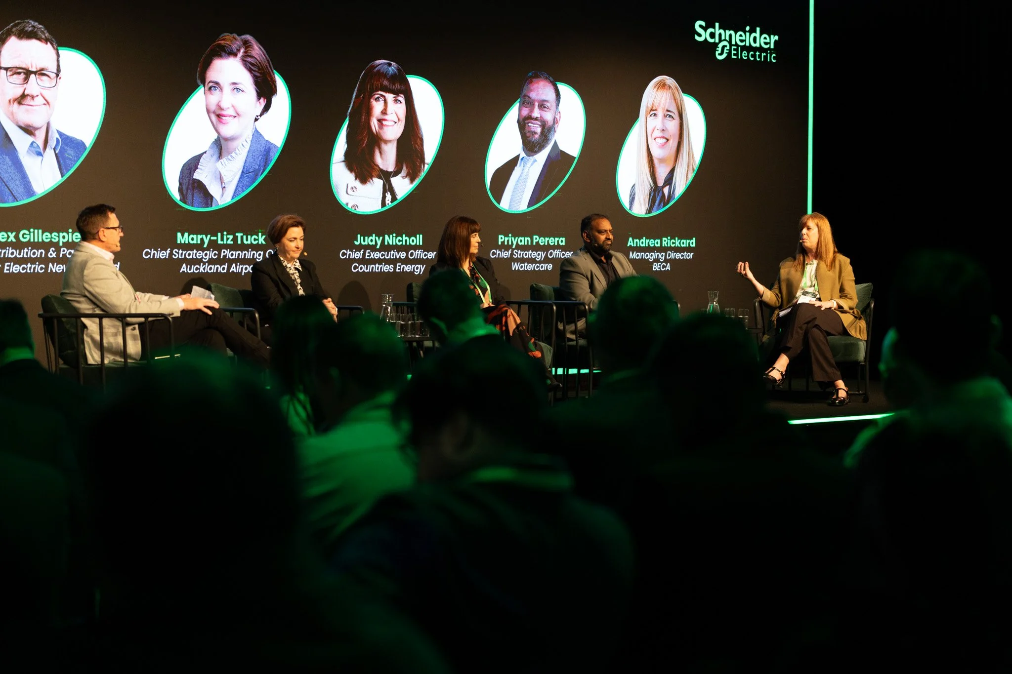 Panel of five professionals speaking at a conference with a large screen behind them displaying images and names, including Alex Gillespie, Mary-Liz Tuck, Judy Nicholl, Priyan Perera, and Andrea Rickard, with the Schneider Electric logo.