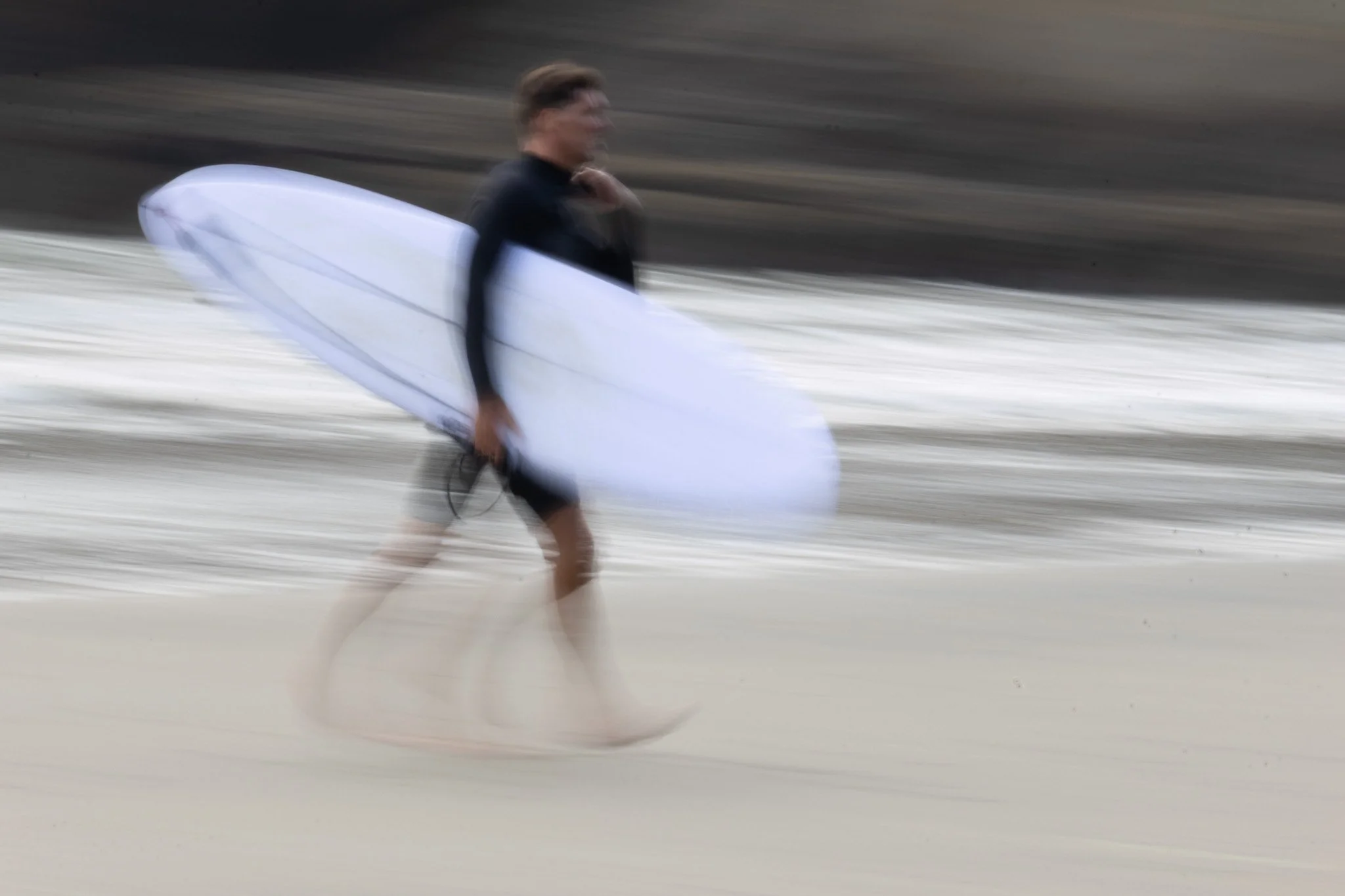 Blurred image of a person walking on the beach carrying a white surfboard.