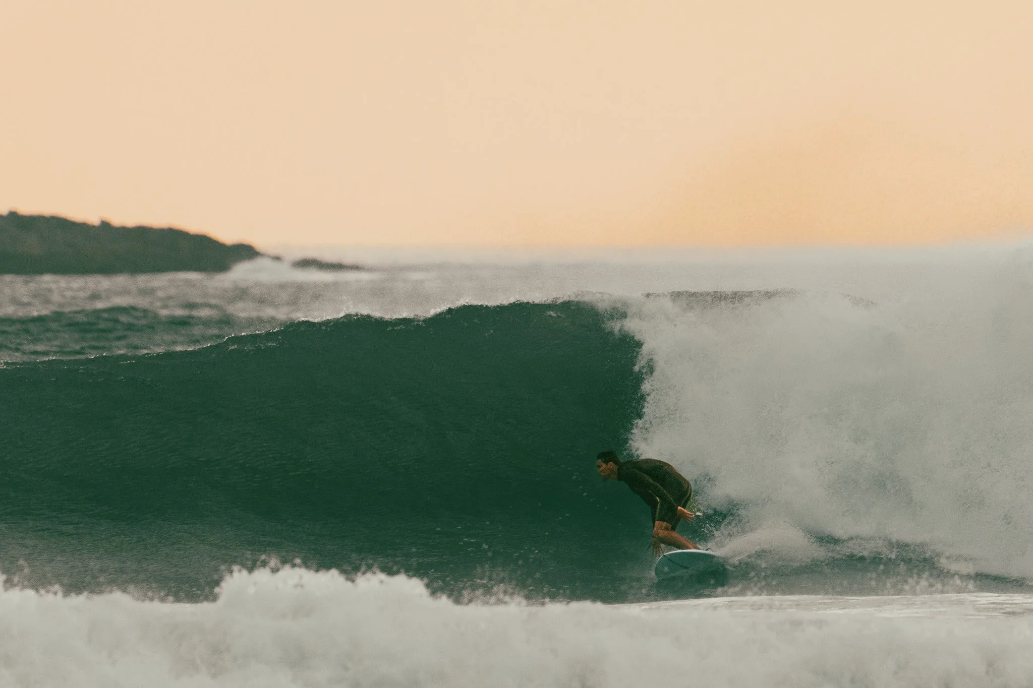 Surfer riding a wave at sunset with distant land in the background.