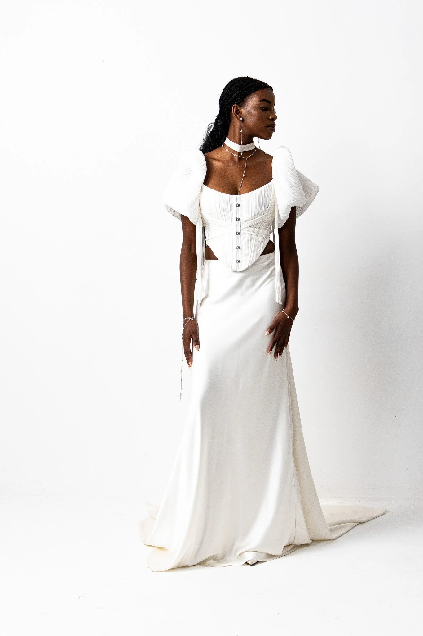 A woman with dark skin wearing a white, elegant, floor-length gown with puffed sleeves, accessorized with layered necklaces and long earrings, standing against a plain white background.