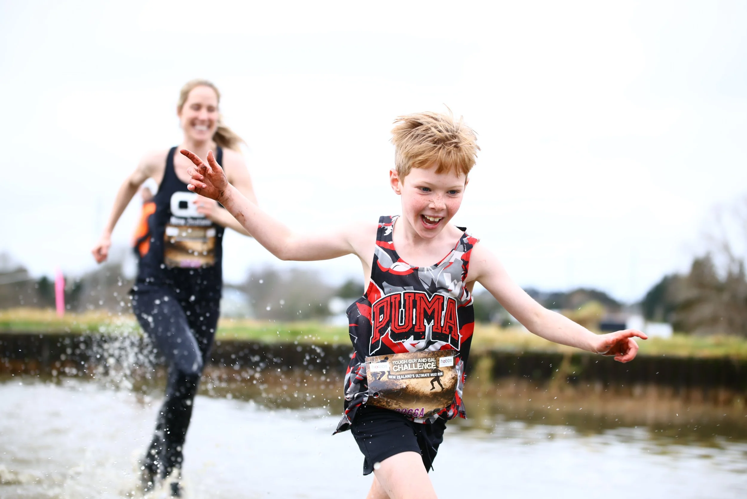 A young boy and a woman running through a shallow muddy water obstacle course during a mud run event, smiling and splashing water.