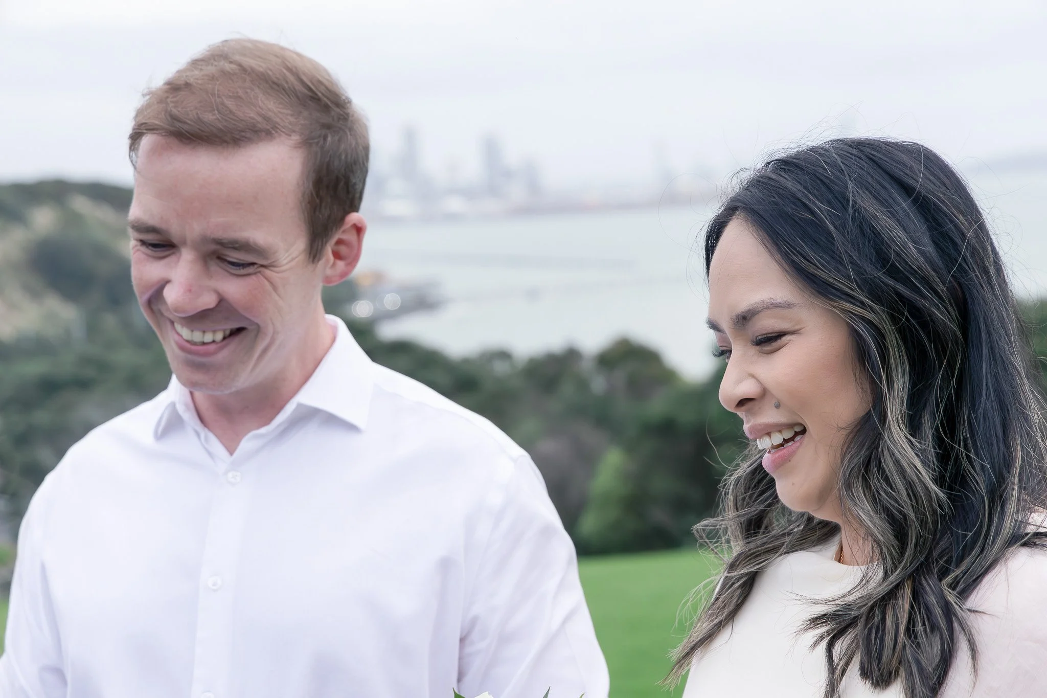 A man and woman smiling outdoors on a cloudy day, with greenery and industrial structures in the background.