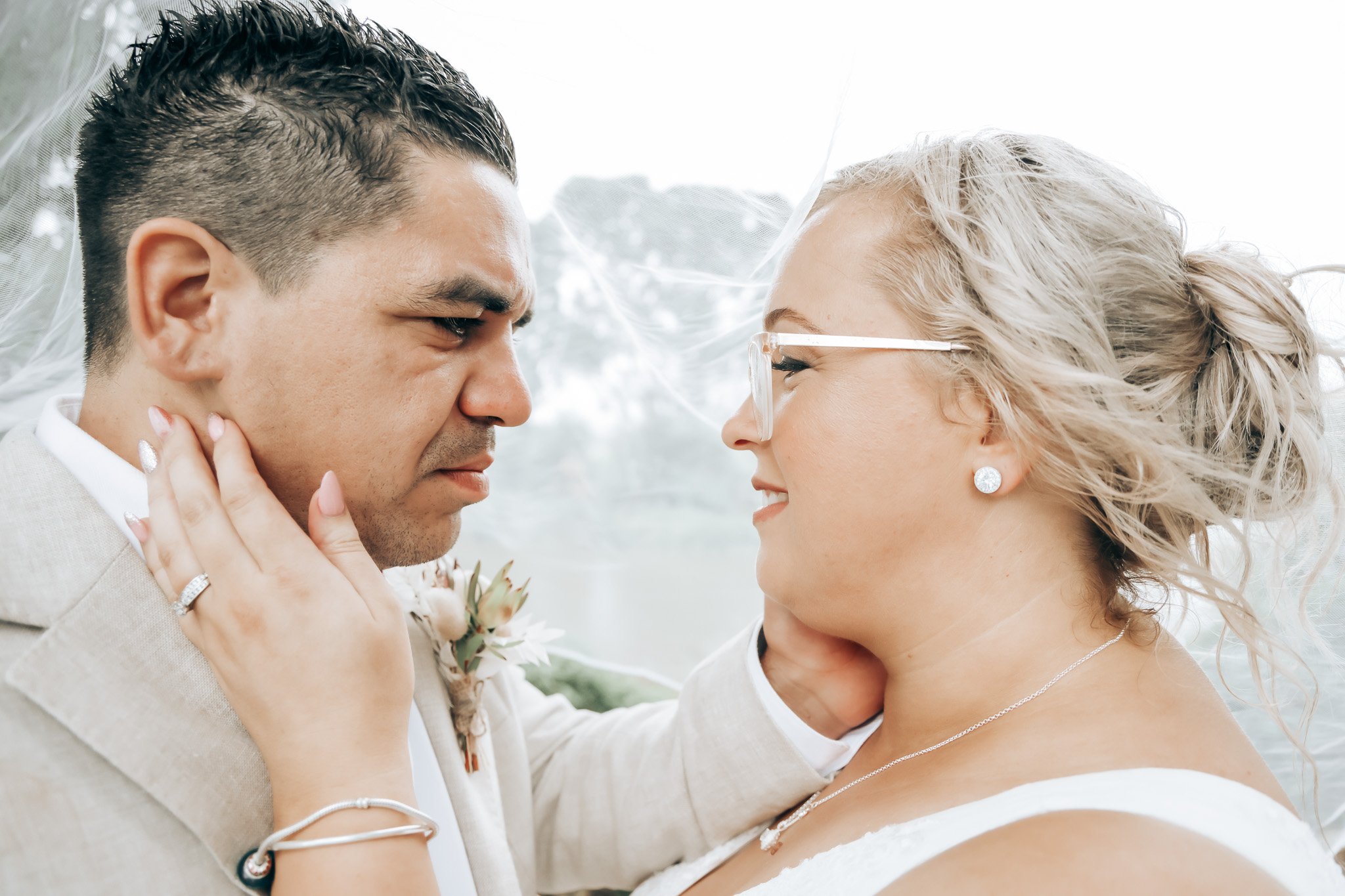 A groom and bride face each other closely, smiling, with their foreheads and noses touching, outdoors with natural light.