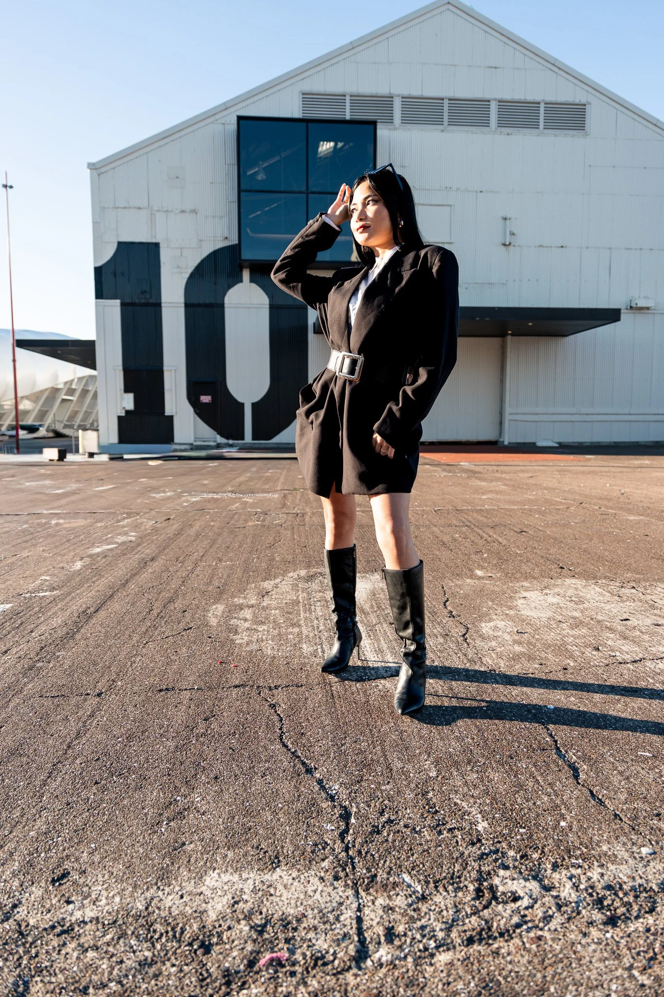 A woman in a black blazer and high-heeled boots standing in front of an industrial building with large windows and a black and white abstract design on the wall, on a cracked asphalt surface under a clear blue sky.