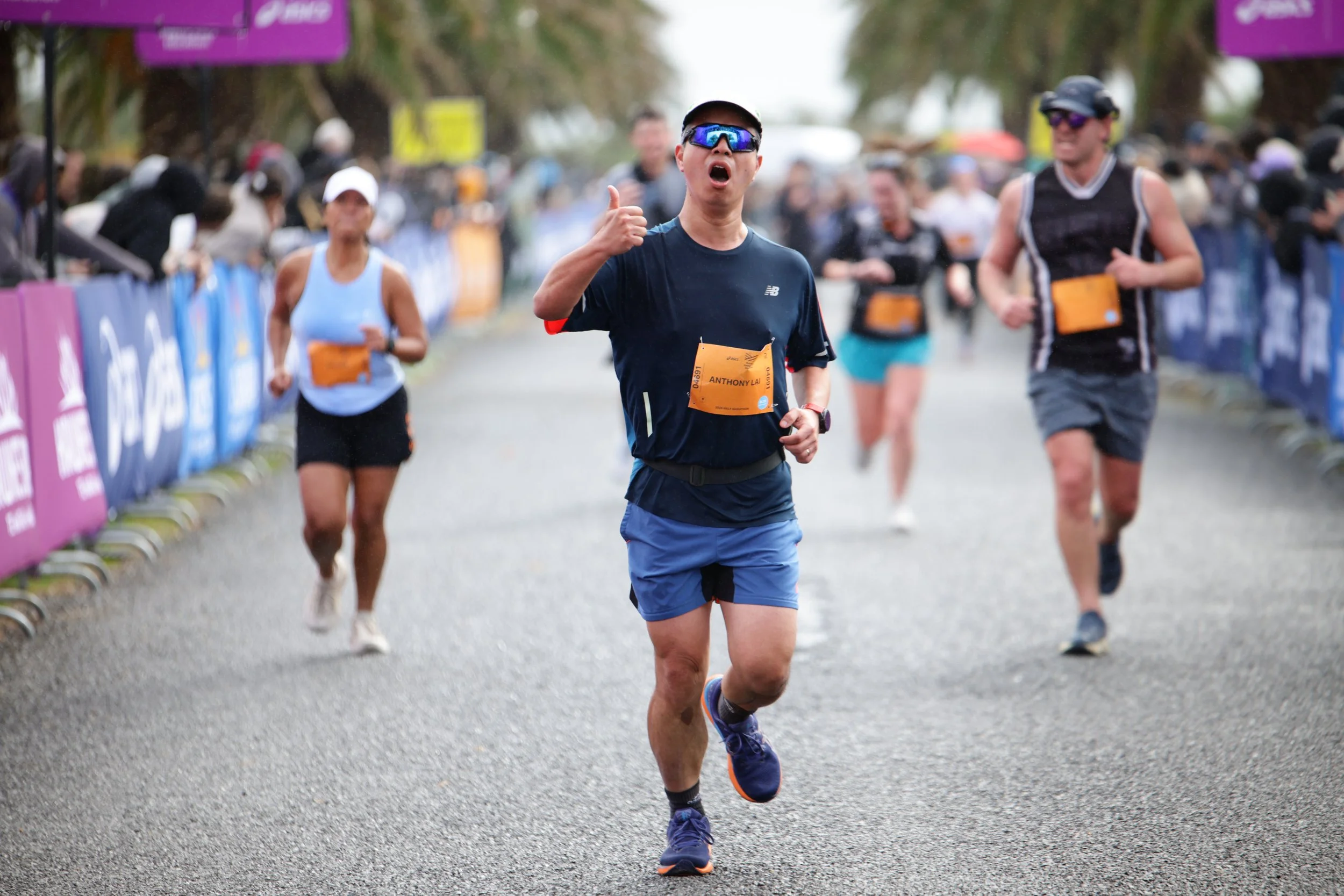 A man wearing sunglasses, a black shirt, and blue shorts running in a race, giving a thumbs-up gesture with a crowd of runners and spectators in the background.