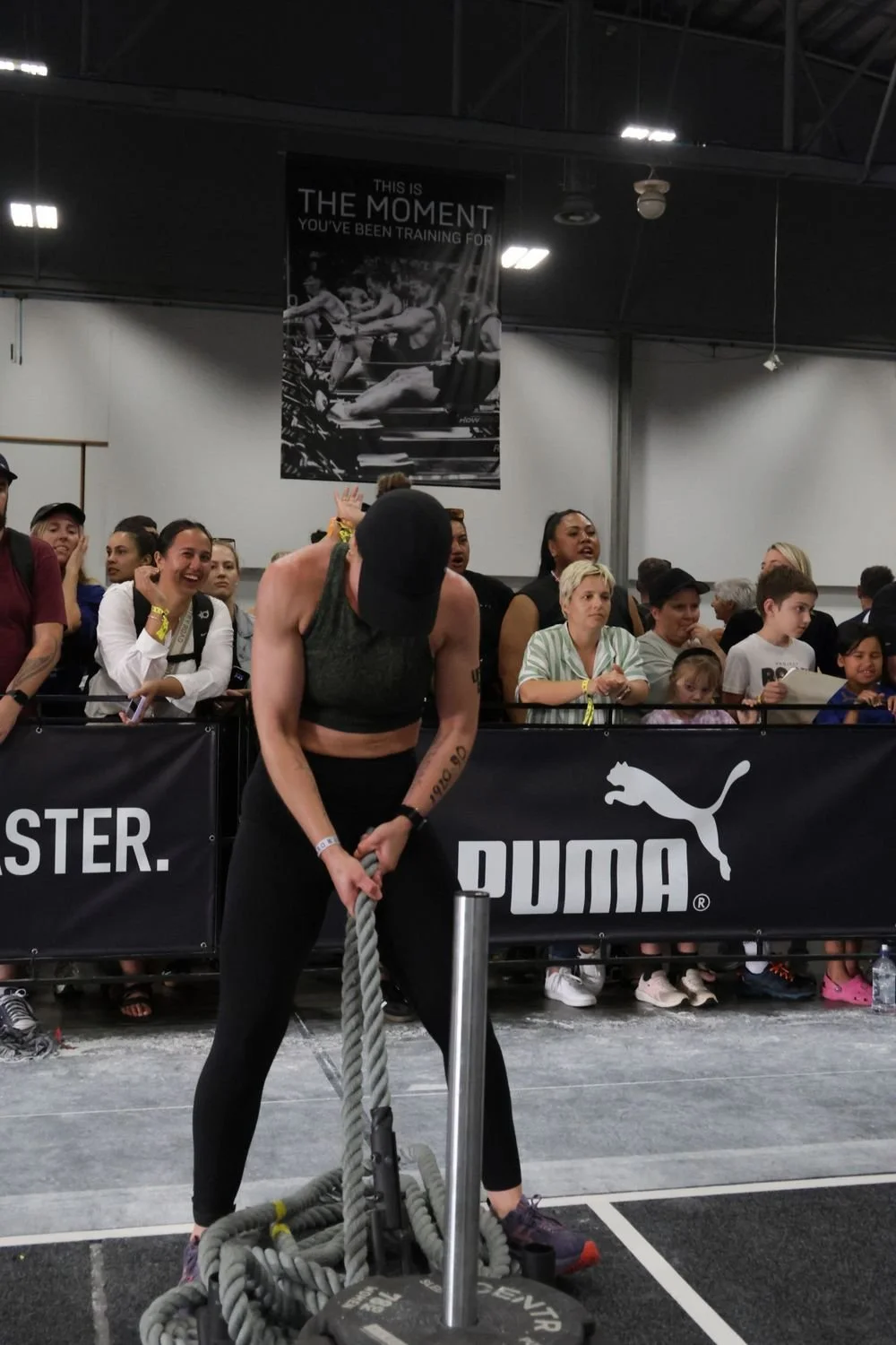 A woman in athletic clothing, wearing a black cap, pulling on a battle rope at a fitness event, with a cheering crowd behind her and a black Puma banner.