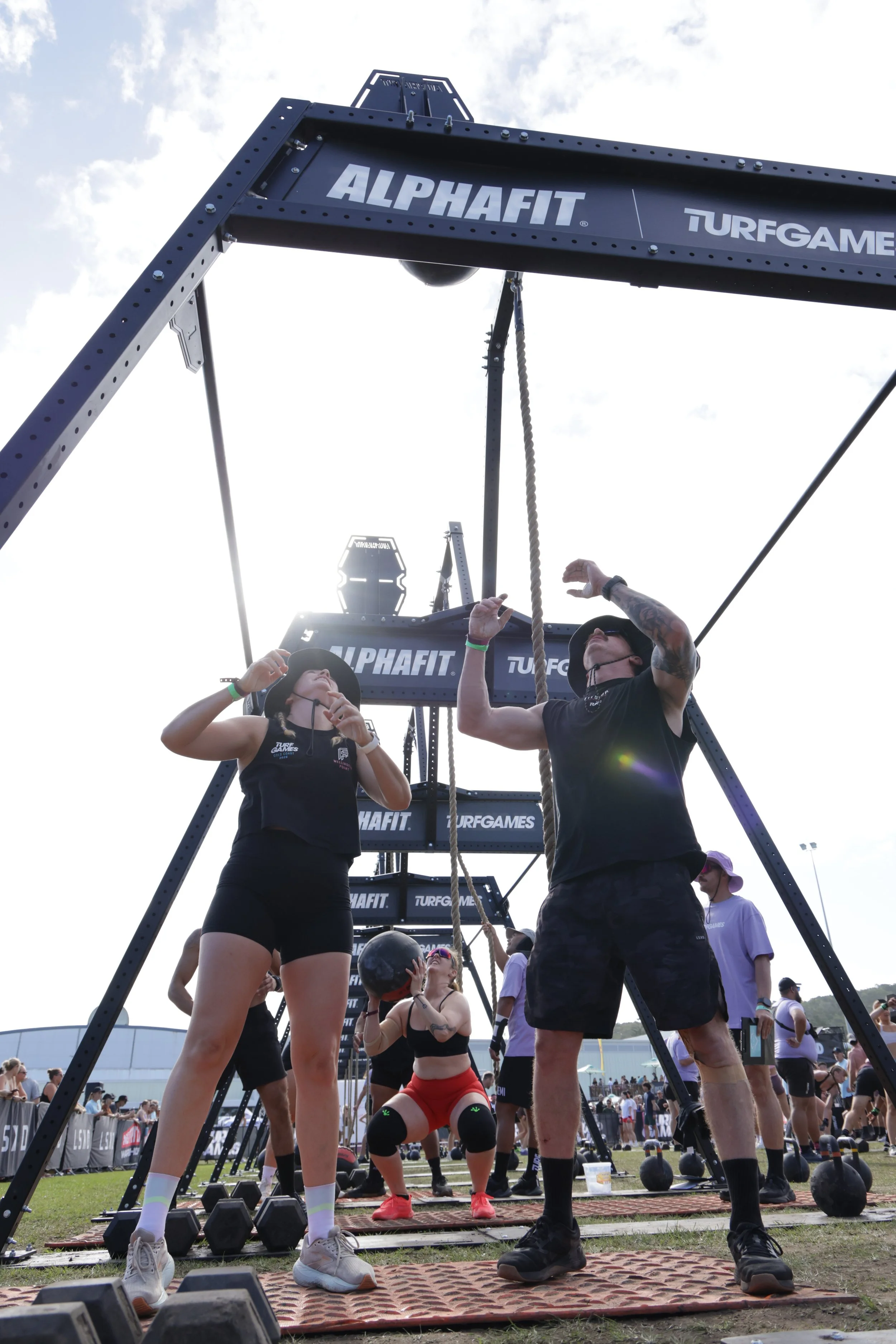 People preparing for a fitness event or workout outdoors, standing on rubber mats with weights and kettlebells, under a large structure with banners labeled 'ALPHAFIT' and 'TURFGAMES'.