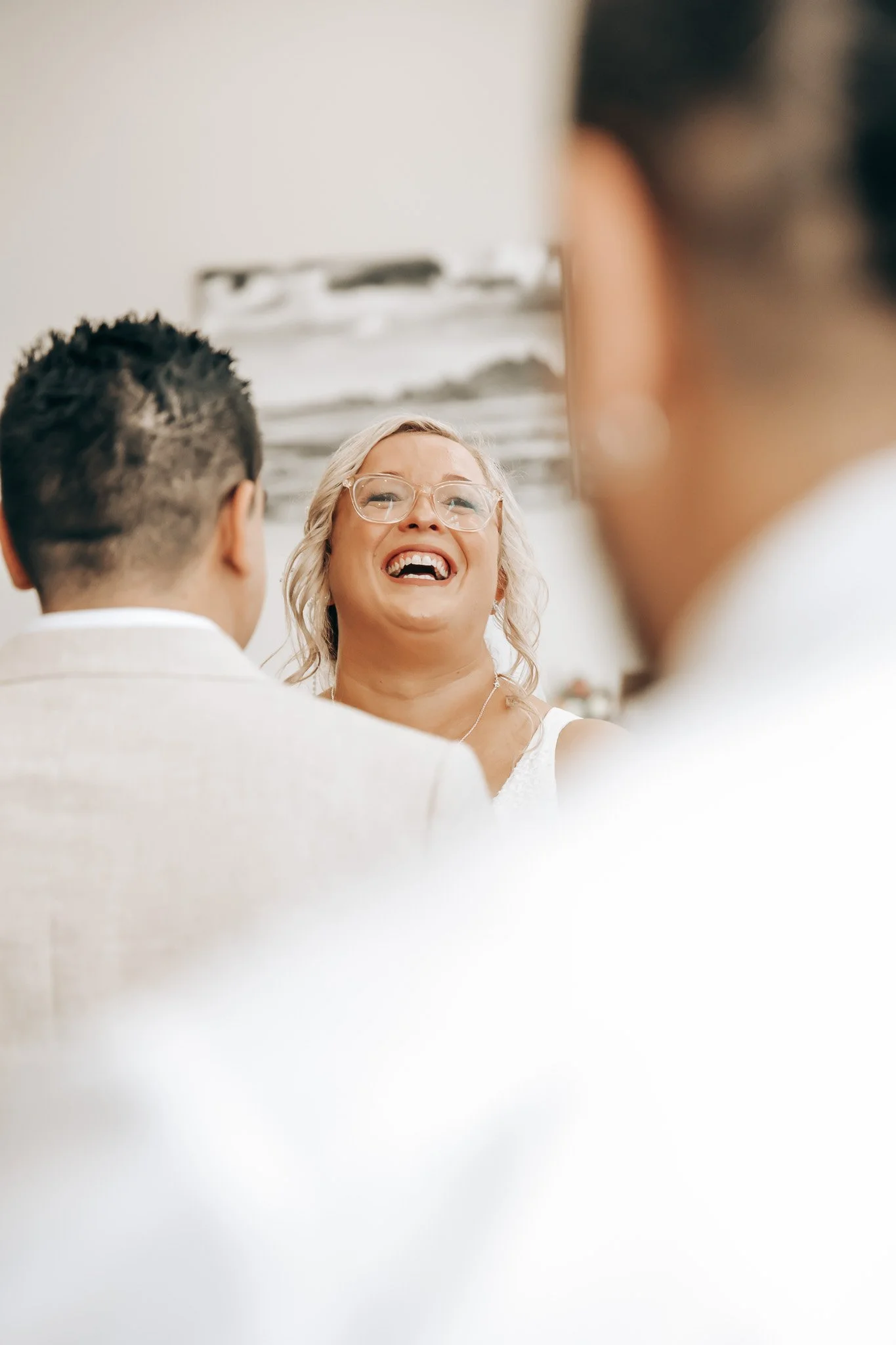 A happy woman with blonde hair and glasses smiling while looking at two men, one with dark hair seen from the back and the other partly visible, in a bright room with abstract artwork in the background.