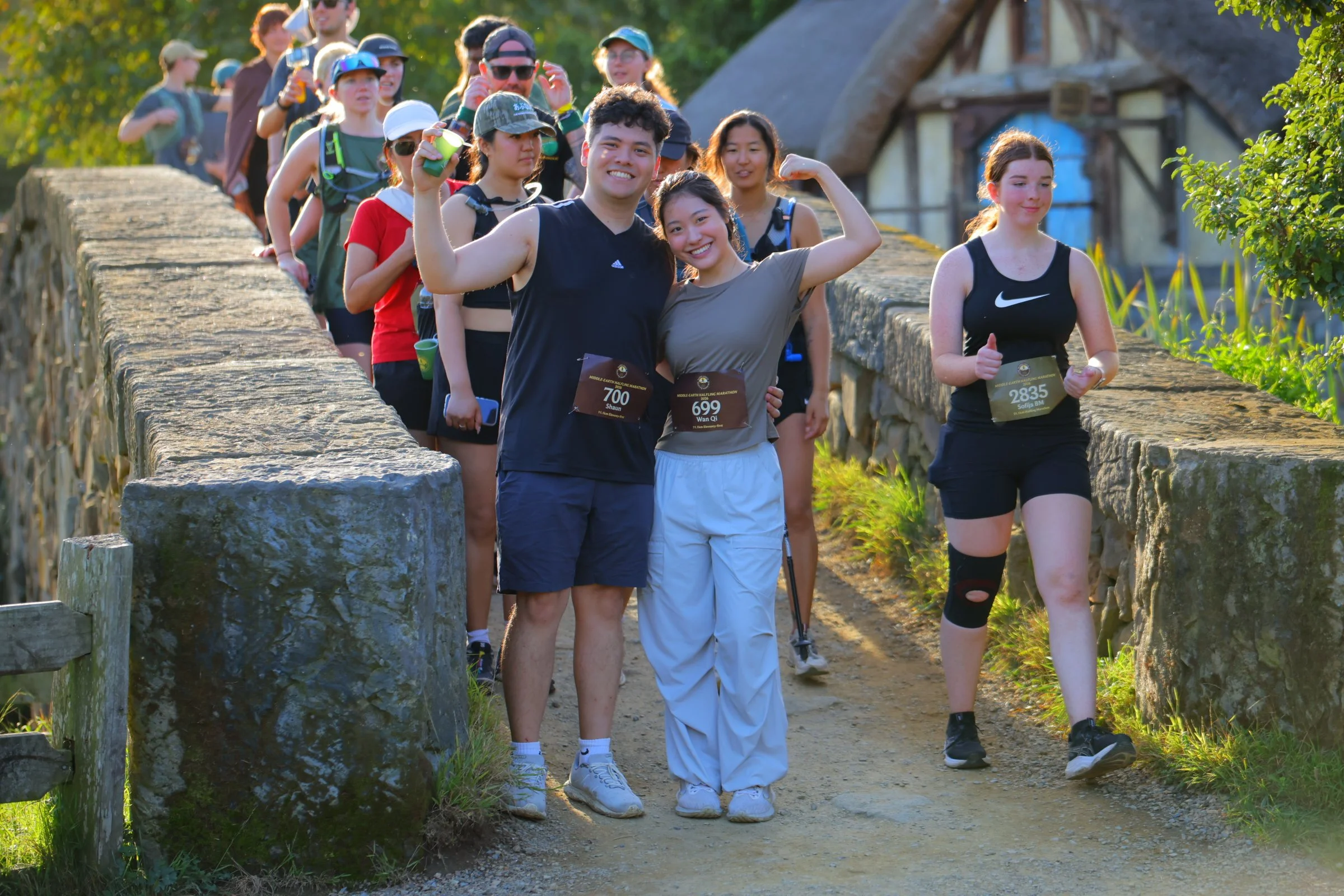 A group of people participating in a marathon or race on a trail, with some smiling and flexing their muscles, outdoors, with greenery and a stone wall.