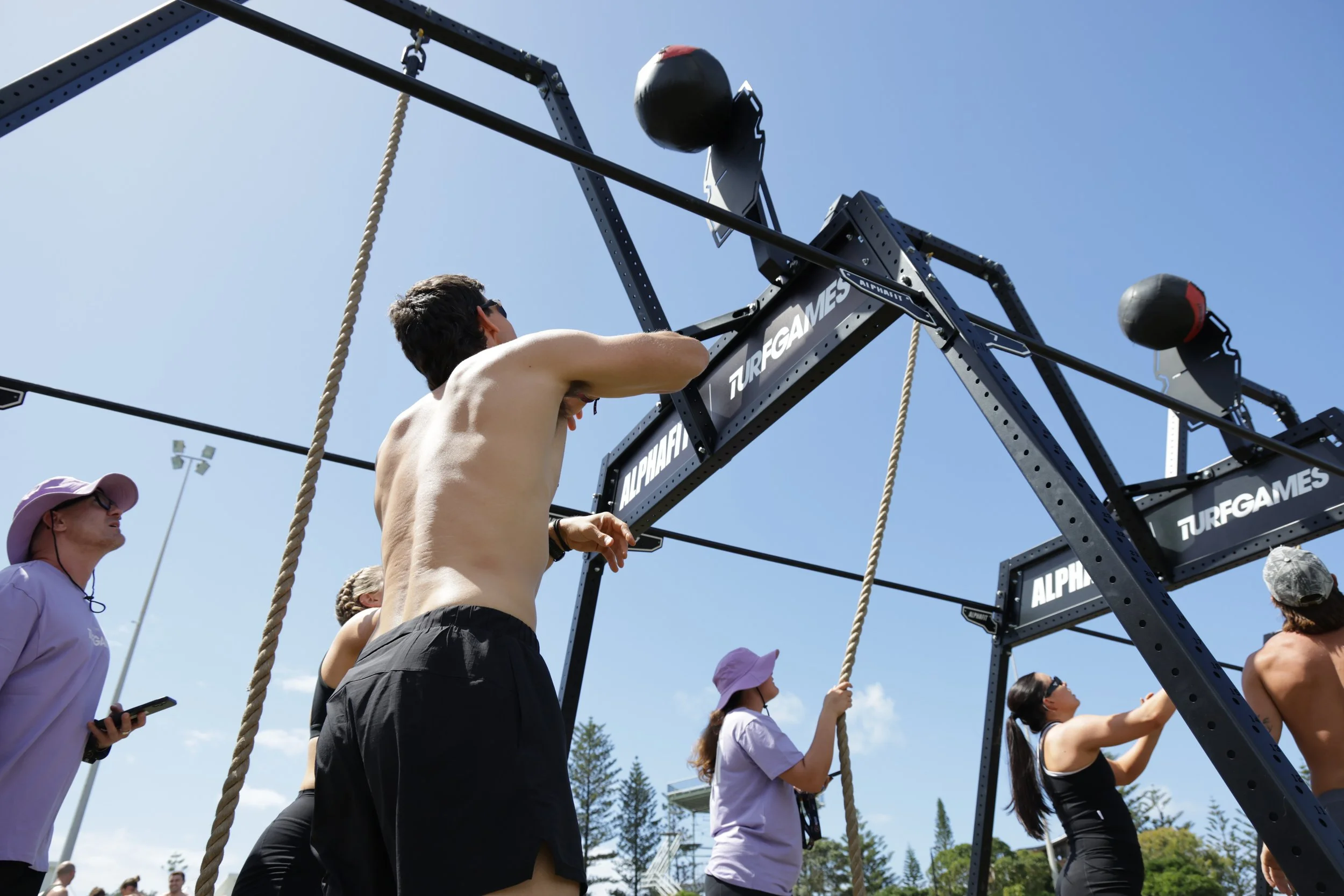 Young man with a muscular build and dark shorts participating in an outdoor fitness competition involving climbing a structured rig with ropes and punching bags, surrounded by spectators under a clear blue sky.