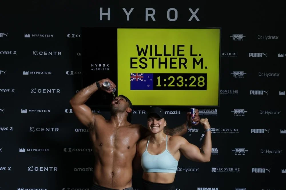 Two athletes, a man and a woman, celebrating at a fitness competition, with the man drinking water and the woman smiling, in front of a black backdrop with brand logos and a yellow scoreboard showing the names Willie L. and Esther M. with a time of 1