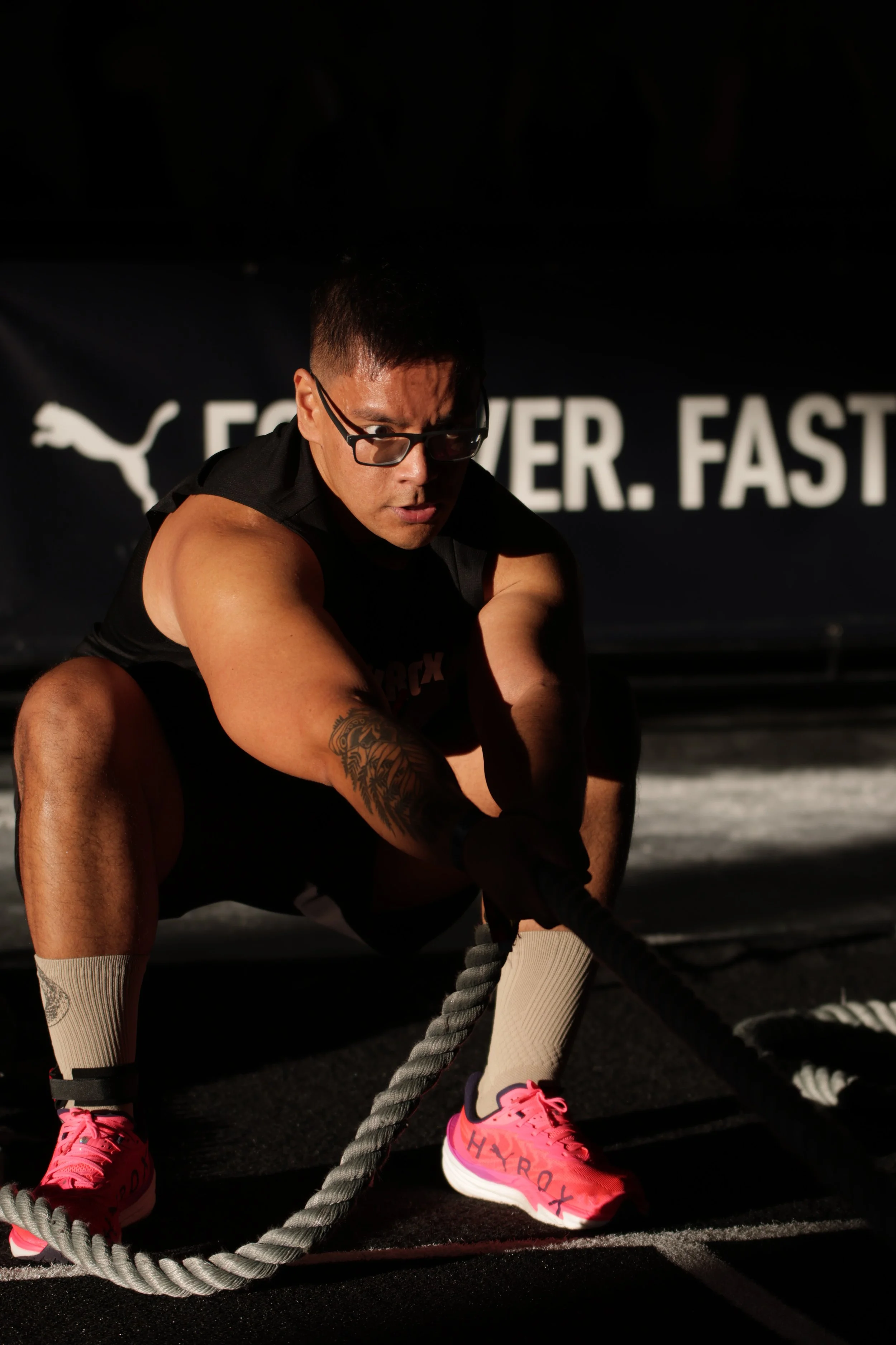 A man in black workout attire, wearing glasses, is sitting in a gym during a workout. He is holding a battle rope and has a tattoo on his upper arm. The background features branding with the Puma logo and the words "FEVER. FAST."