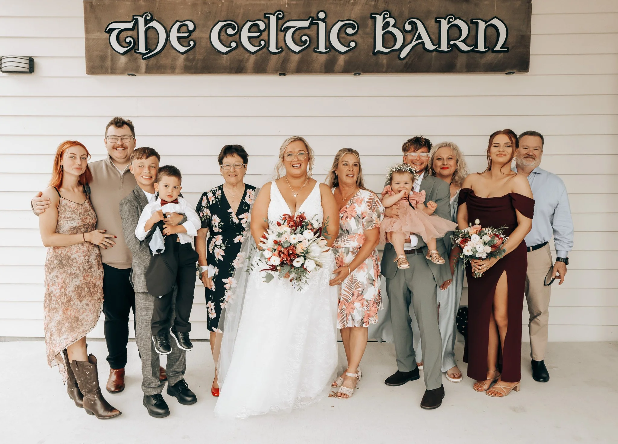 A group of twelve people, including children and adults, standing in front of a sign that reads 'the Celtic BAR' on a white wall. The group includes women, men, and children dressed in formal and casual attire, smiling for a group photo at what appea