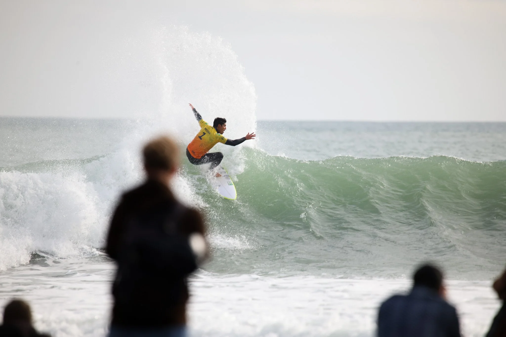 Surfer riding a wave at the beach, with several spectators in the foreground.