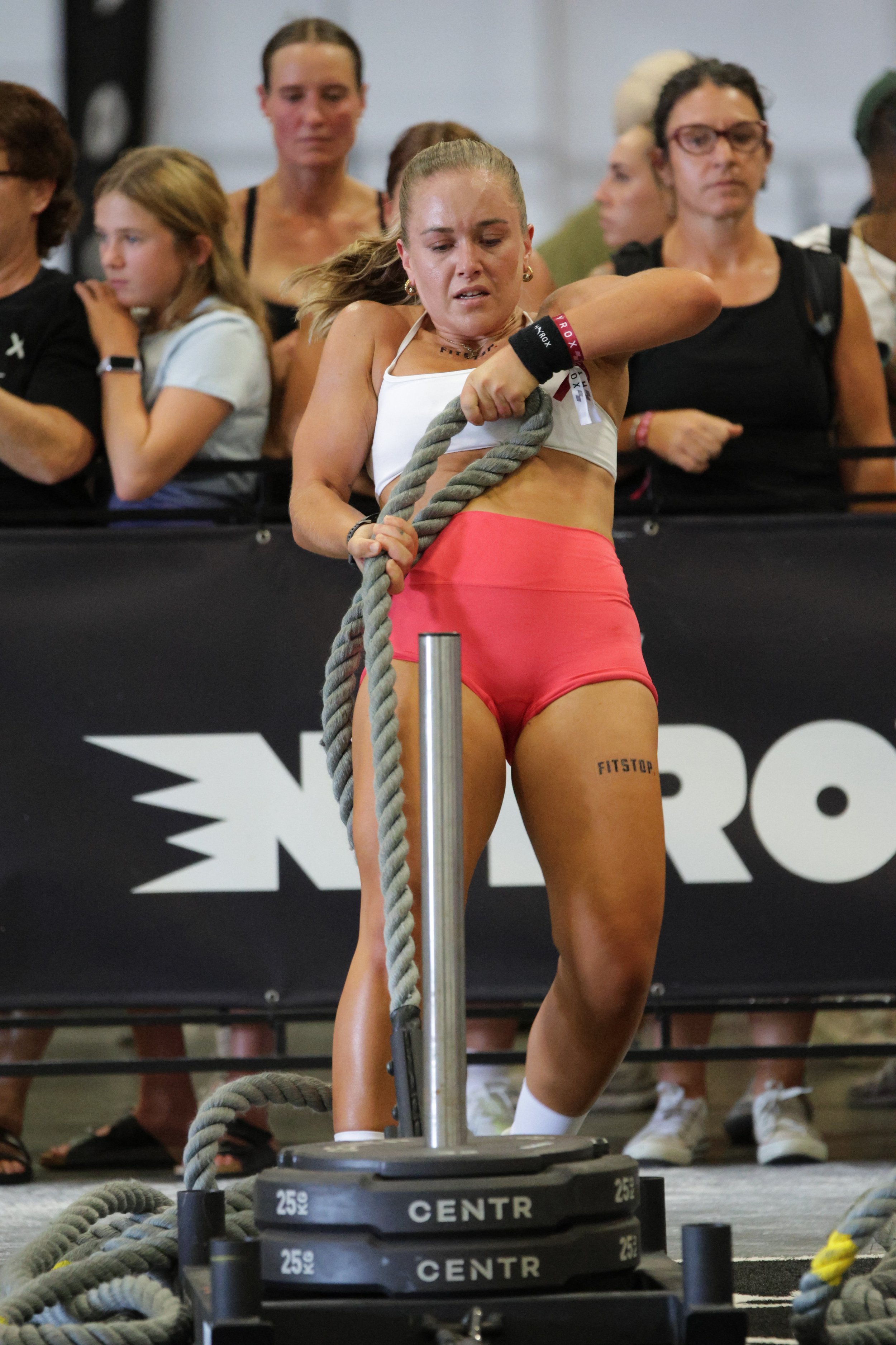 A woman in a white sports bra and pink shorts participating in a fitness competition, pulling on a thick rope. Several spectators stand behind her, watching. The scene is indoors, with fitness equipment and a black barrier in the background.