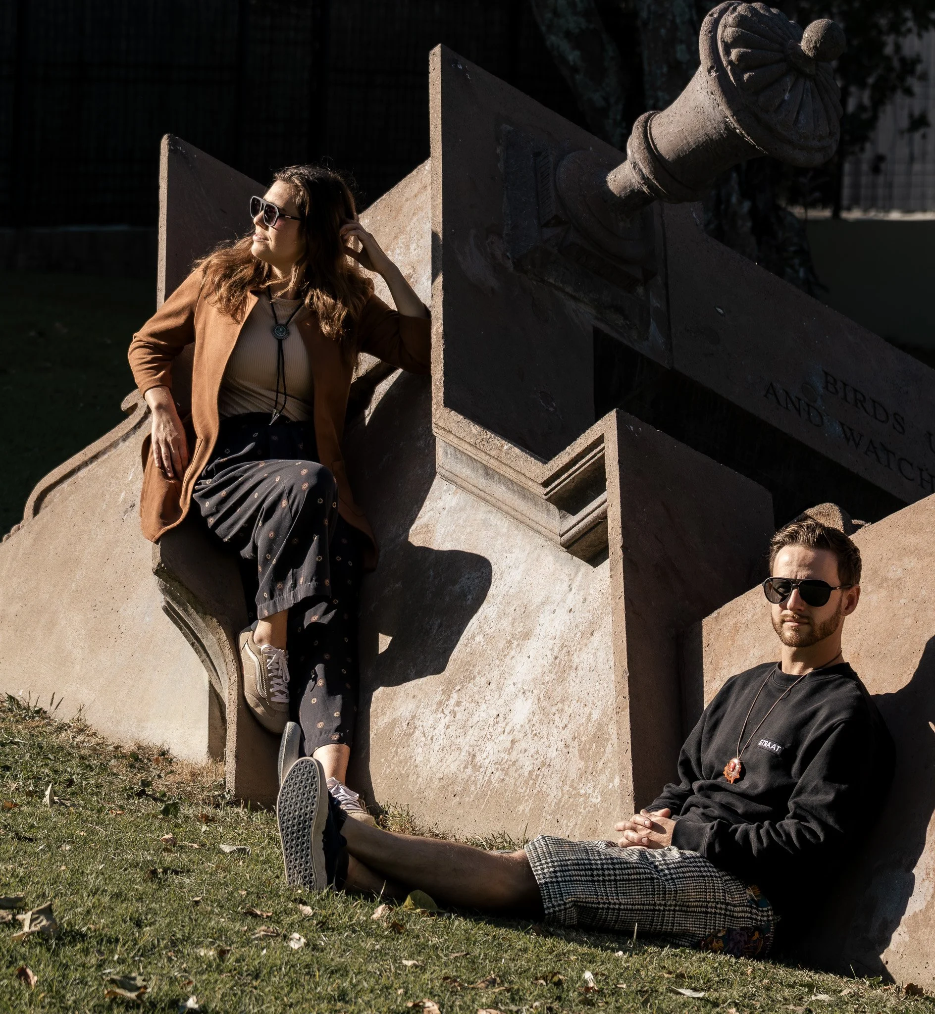 Three young people sitting outdoors on the grass near a stone monument with a decorative fountain spout. The woman on the left wears sunglasses, a brown jacket, and dark patterned pants, and has long wavy hair. The man on the right wears sunglasses, 