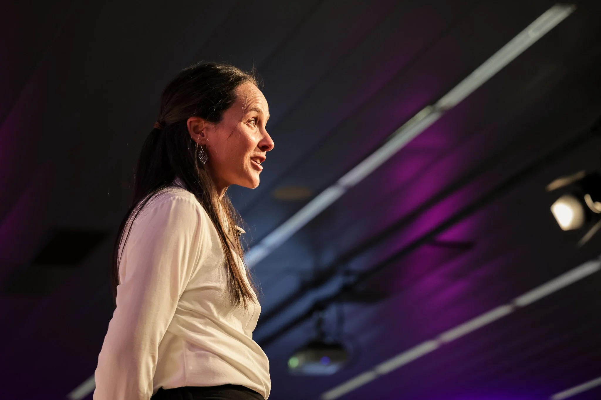 A woman with long dark hair, wearing earrings and a light-colored blouse, is standing in profile against a dark ceiling with purple and white lighting.