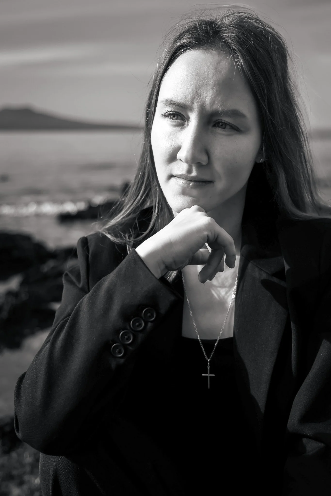 Close-up black and white photo of a young woman with light-colored eyes and long hair, wearing a dark blazer and a cross necklace, outdoors with blurred landscape background.
