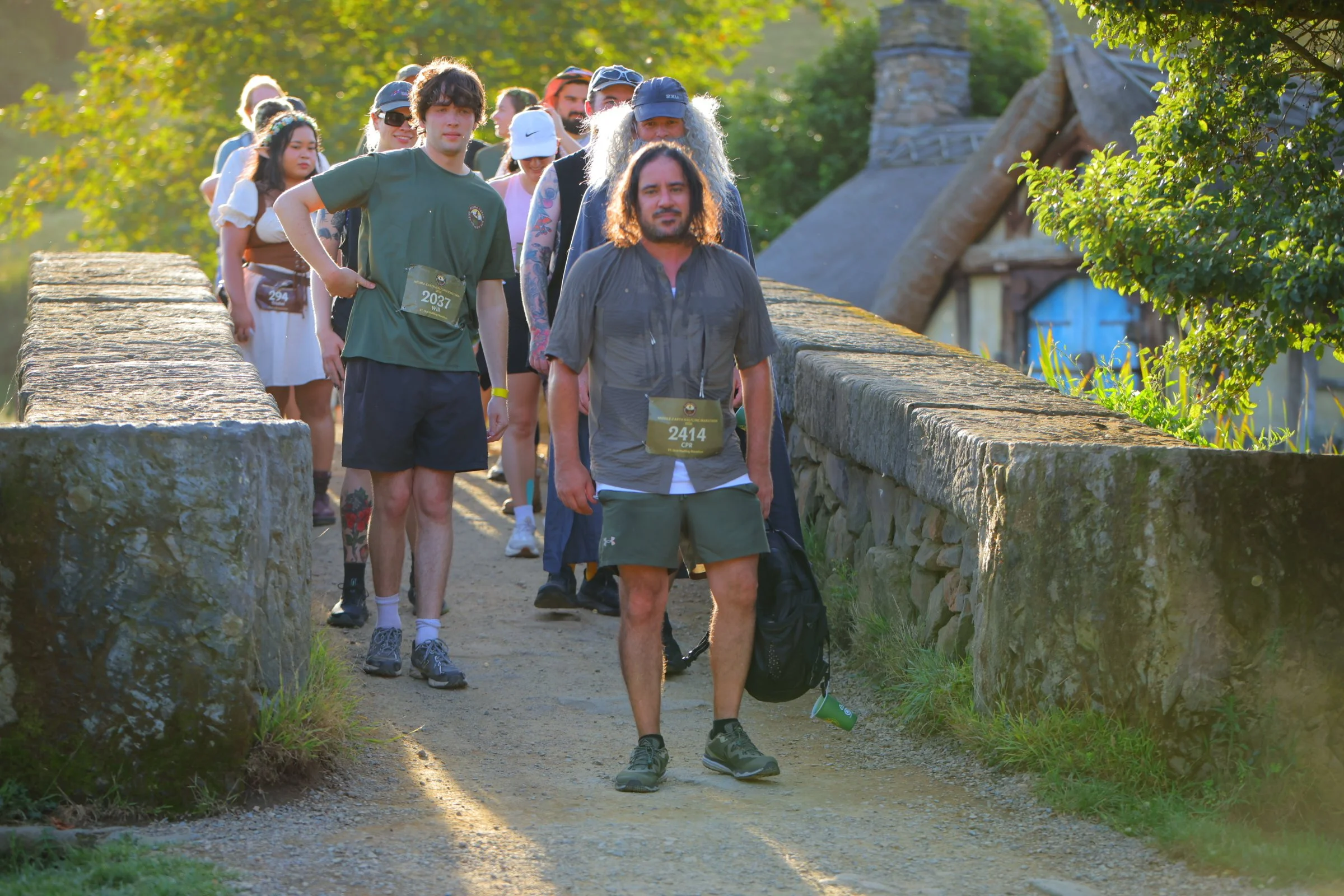 A group of people walking on a stone path in a park or natural setting, with trees and a house in the background, during sunny weather.