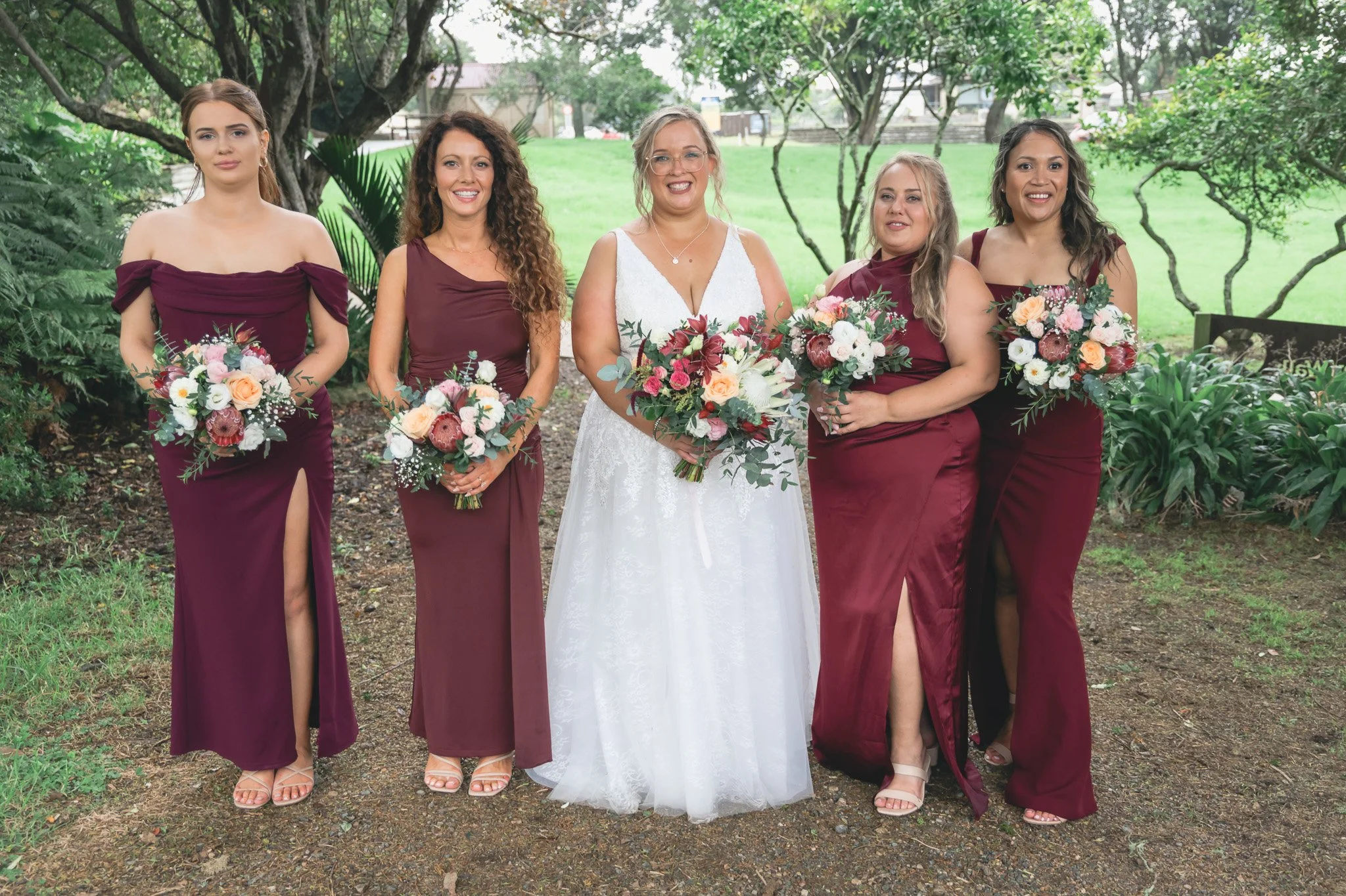 A bride in a white wedding dress standing with five bridesmaids in maroon dresses holding bouquets outdoors in a green park.