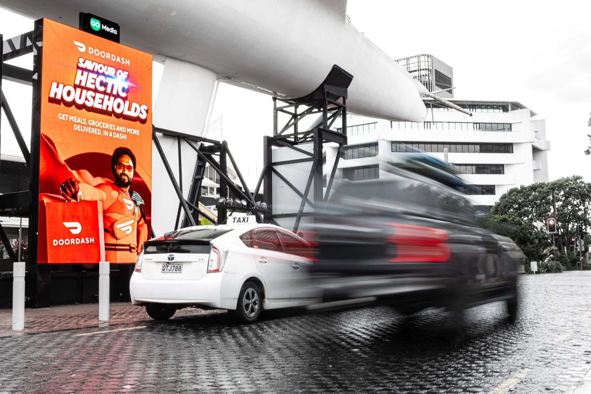A busy urban street scene with a large digital billboard advertising DoorDash, with a driver in motion blur passing by, and modern buildings in the background.