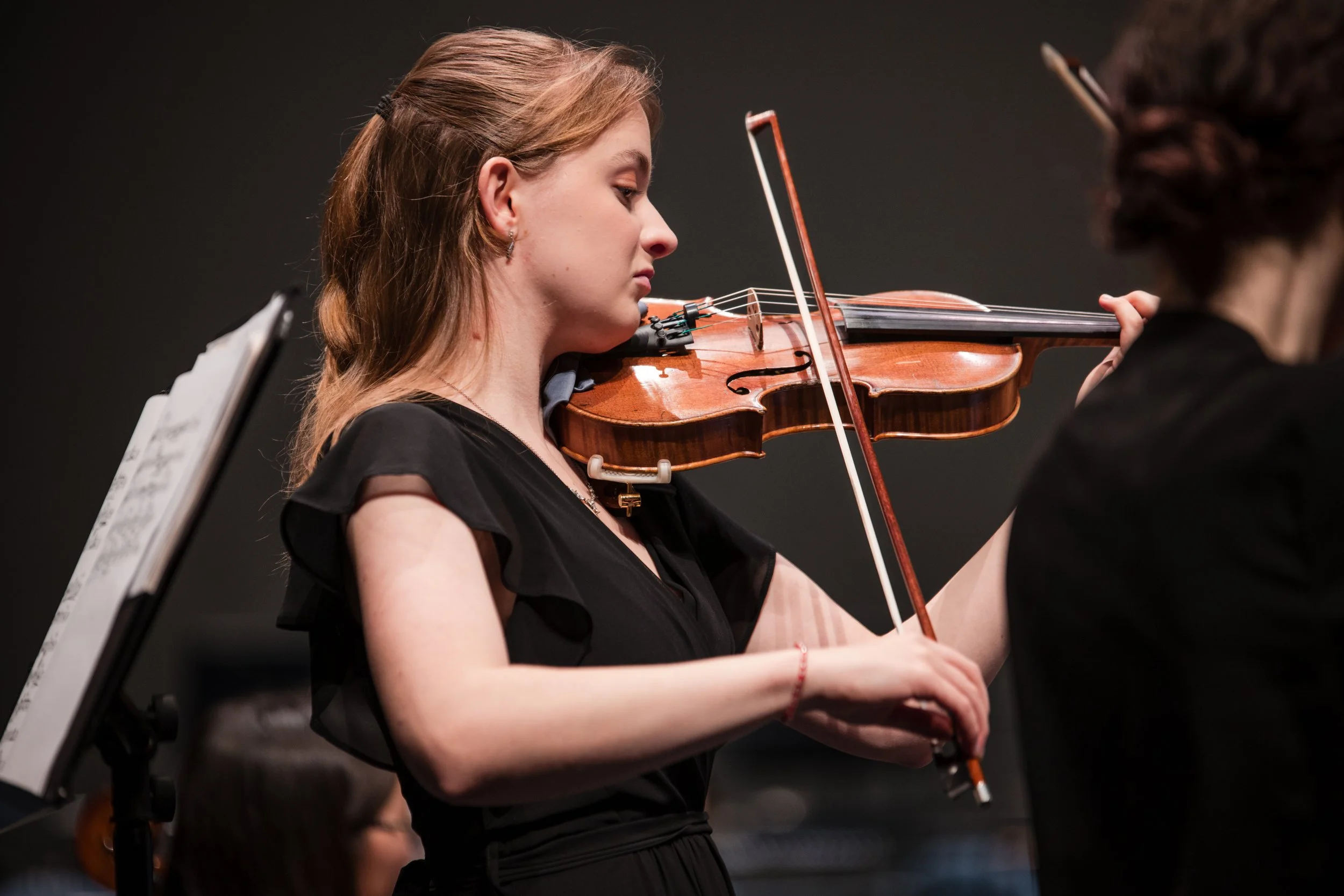 A young woman with light brown hair tied back, playing a violin during a performance or rehearsal, with sheet music on a stand nearby.
