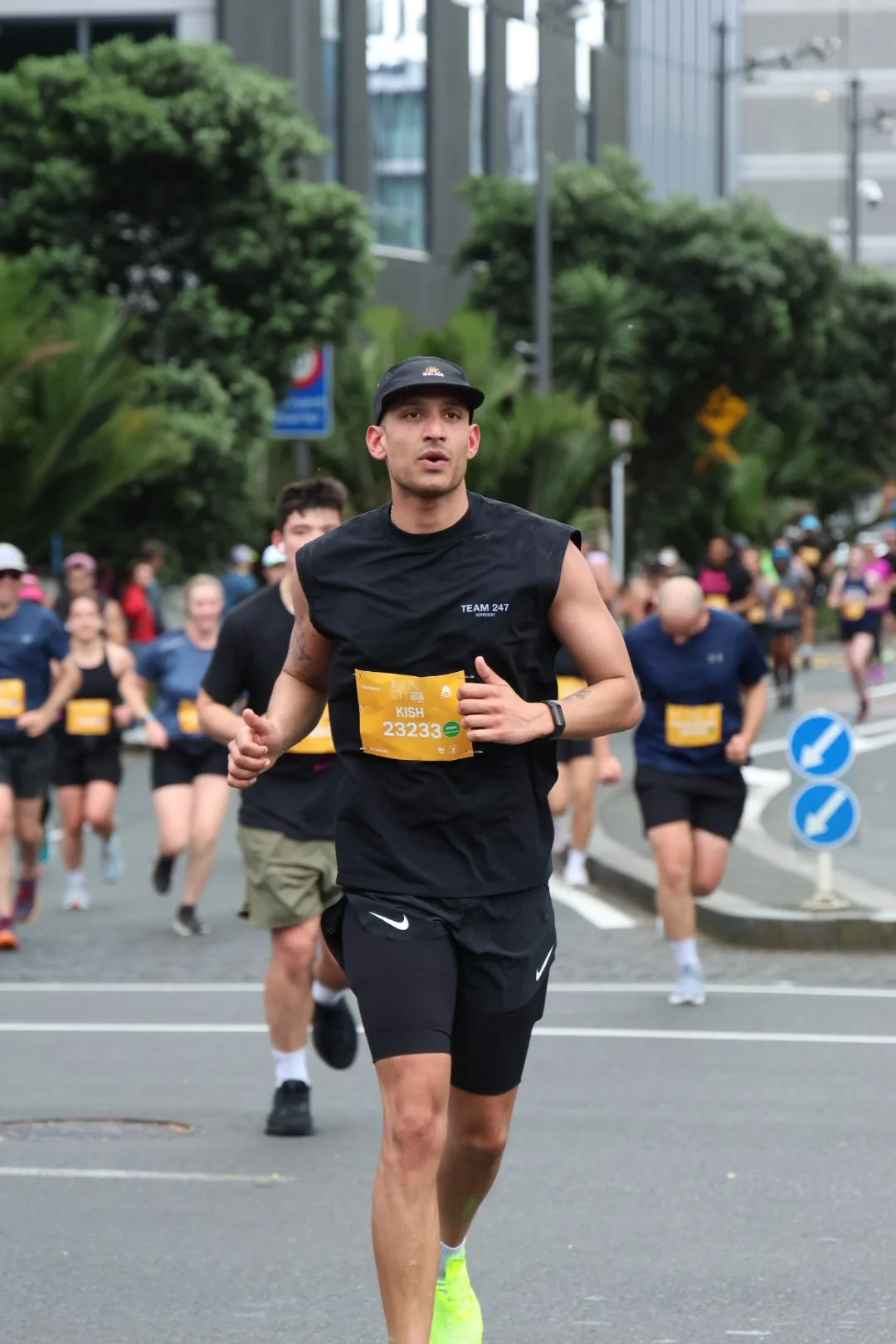 A group of people running in a marathon on city streets, with trees and modern buildings in the background, and a runner in the foreground wearing a black sleeveless shirt, black shorts, a cap, and a yellow race bib.