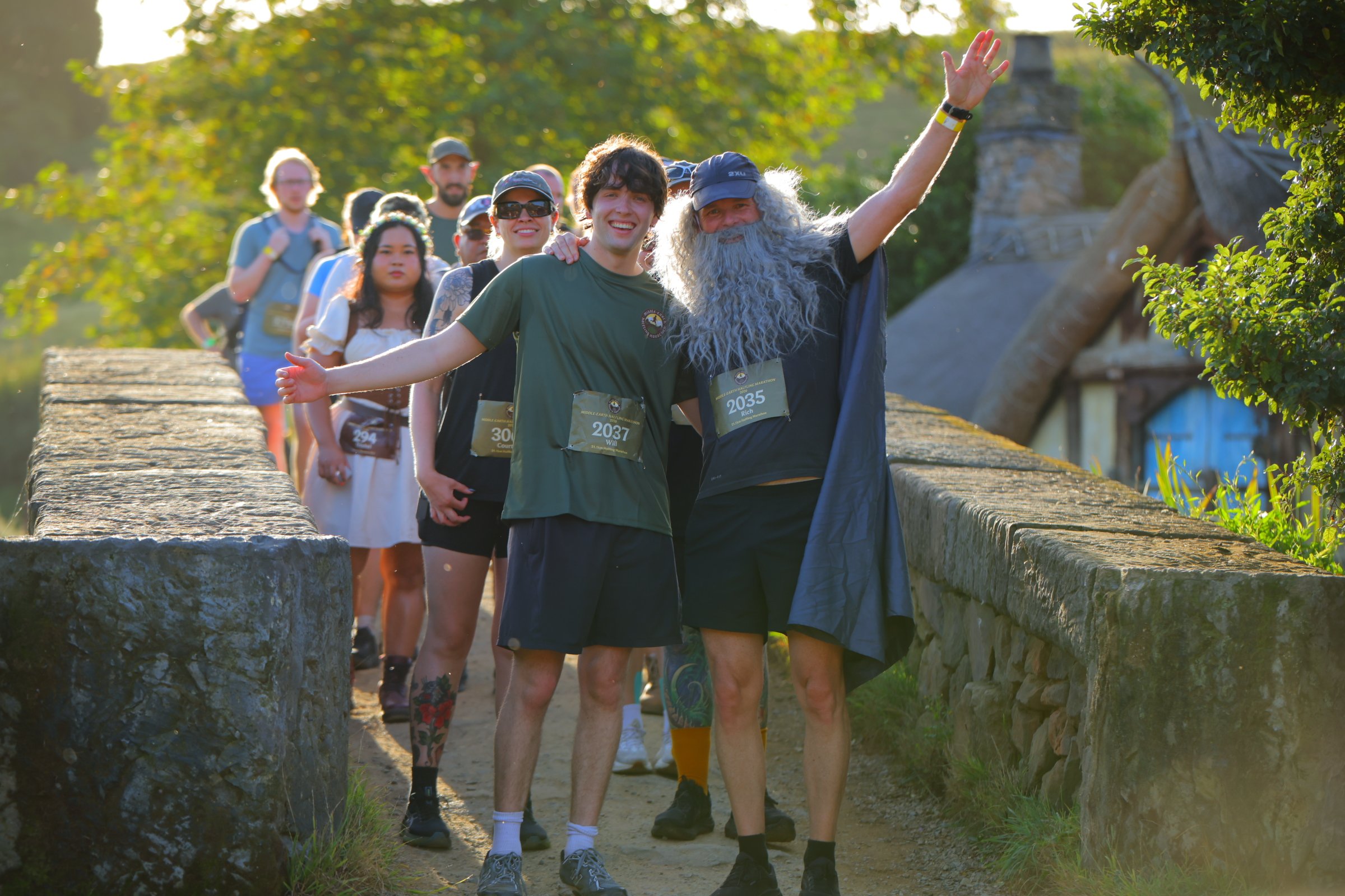 A group of people participating in a marathon or walkathon outdoors, led by a man with a long gray beard dressed as a wizard or mythical character, smiling and waving at the camera. The group is walking on a stone pathway surrounded by trees and old 