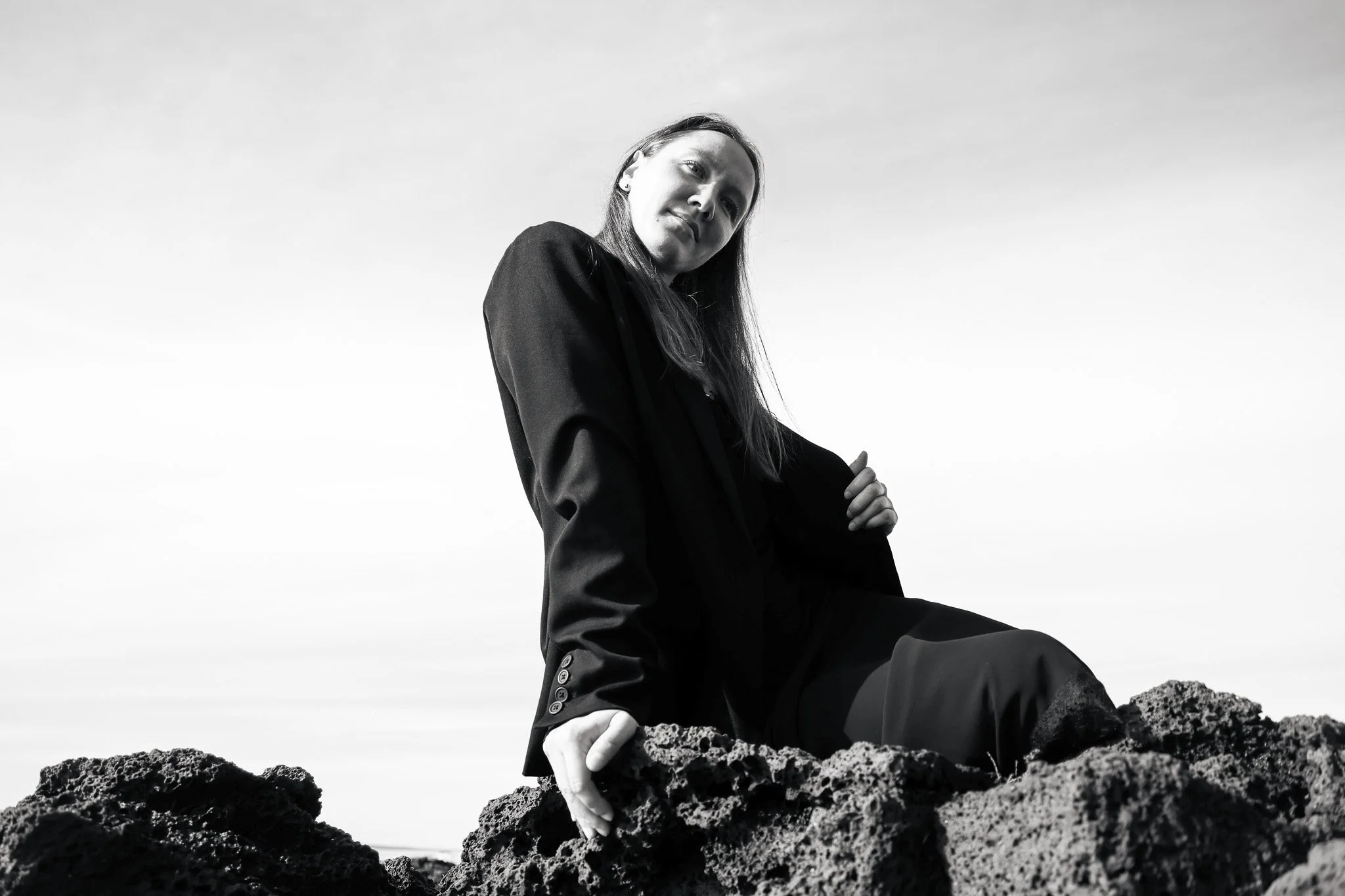 Black and white photo of a woman sitting on volcanic rocks, wearing a dark jacket and pants, with her arm resting on her knee, looking thoughtfully into the distance against a cloudy sky.