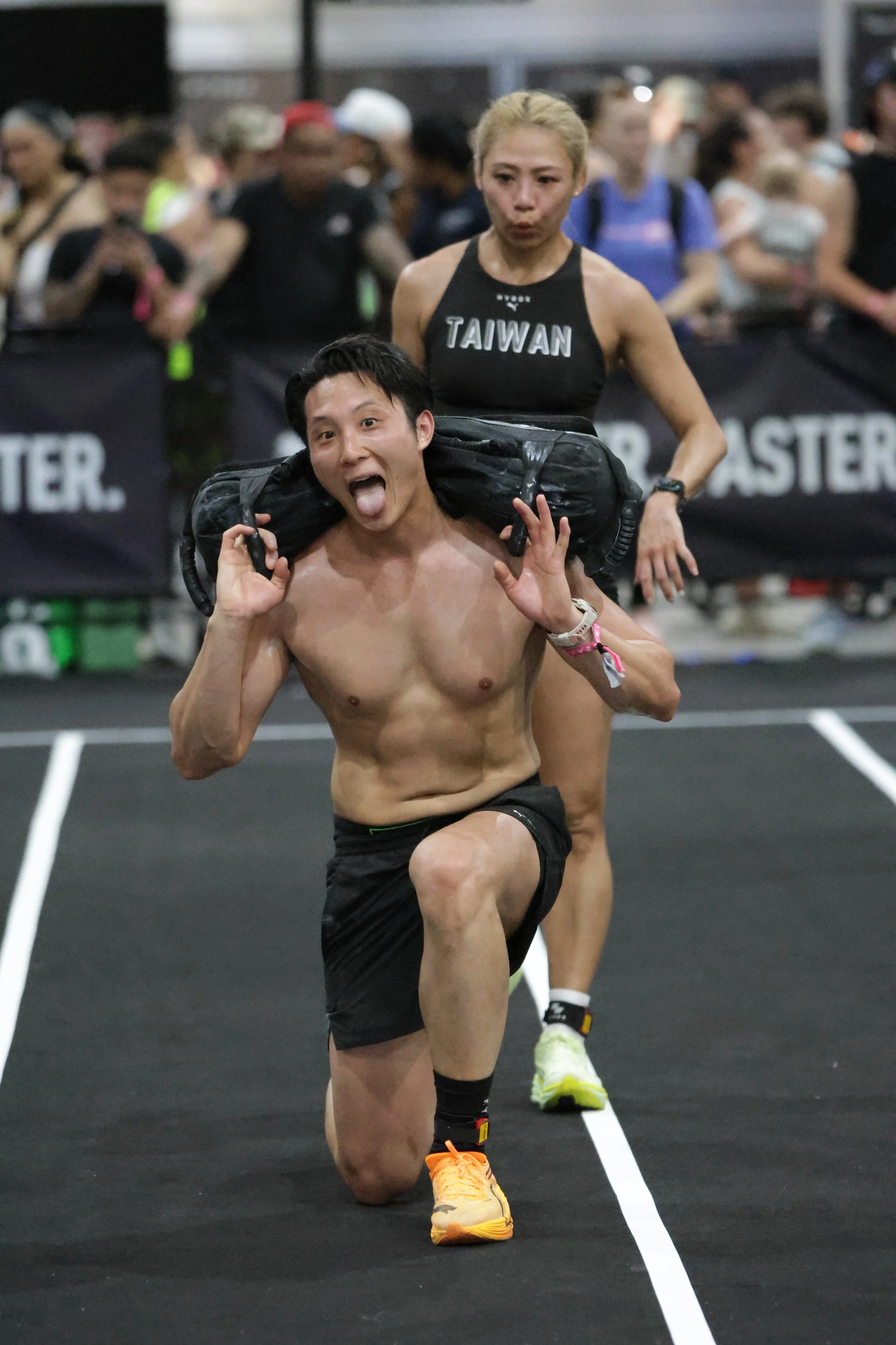 A shirtless man wearing black shorts and yellow running shoes runs with a backpack on his shoulders, followed by a woman wearing a black tank top with 'Taiwan' written on it, in a crowded indoor running event.