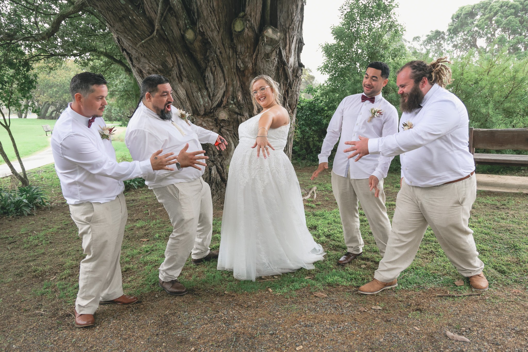 A bride and five groomsmen having fun and dancing outdoors under a large tree during a wedding celebration.