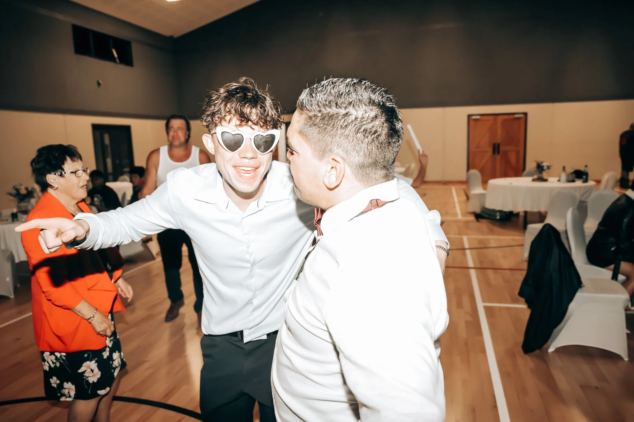 Two young men are dancing and laughing in a ballroom, one wearing heart-shaped sunglasses and a white shirt, the other in a white shirt with a brown collar, surrounded by other guests and round tables.