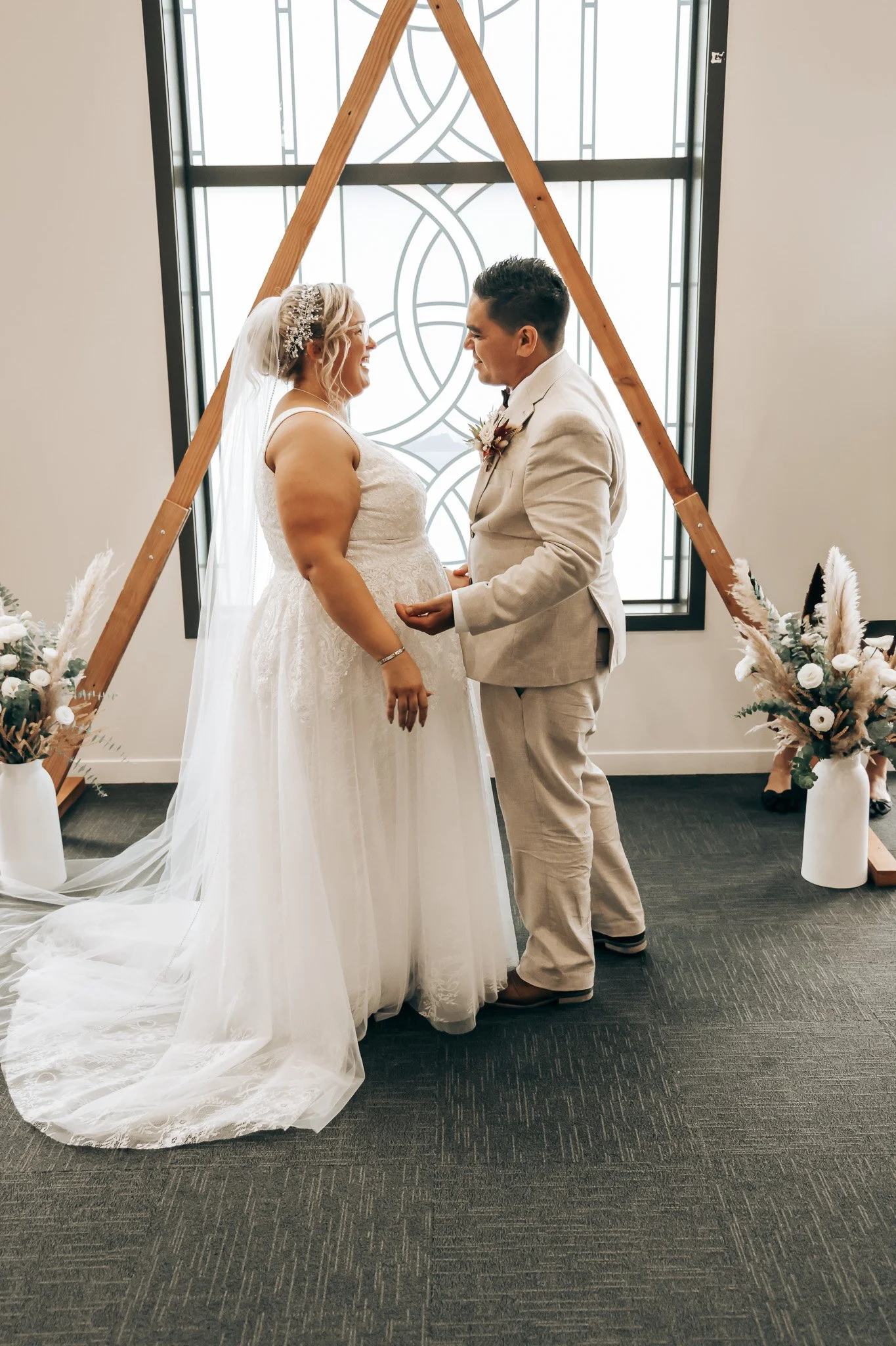 A bride and groom standing face to face, smiling and holding hands during a wedding ceremony in front of a decorative window, with flower arrangements on either side.
