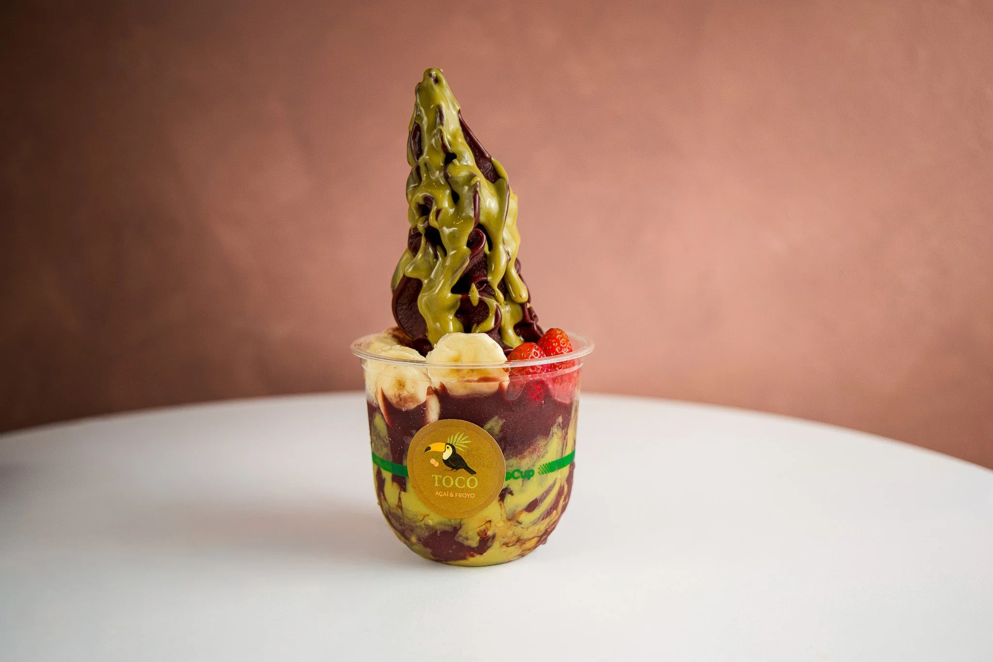 A colorful frozen dessert in a clear plastic cup with a toucan logo, topped with bananas, strawberries, chocolate, and a green sauce.