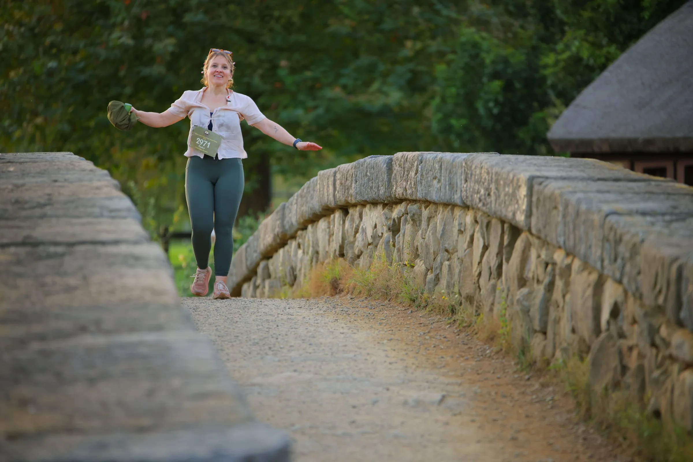 A woman with blonde hair, wearing athletic clothing and a race bib number 2971, balancing on a stone wall in a park or nature setting during late afternoon or early evening. She is smiling, holding a green hat in her left hand, with a backdrop of gre