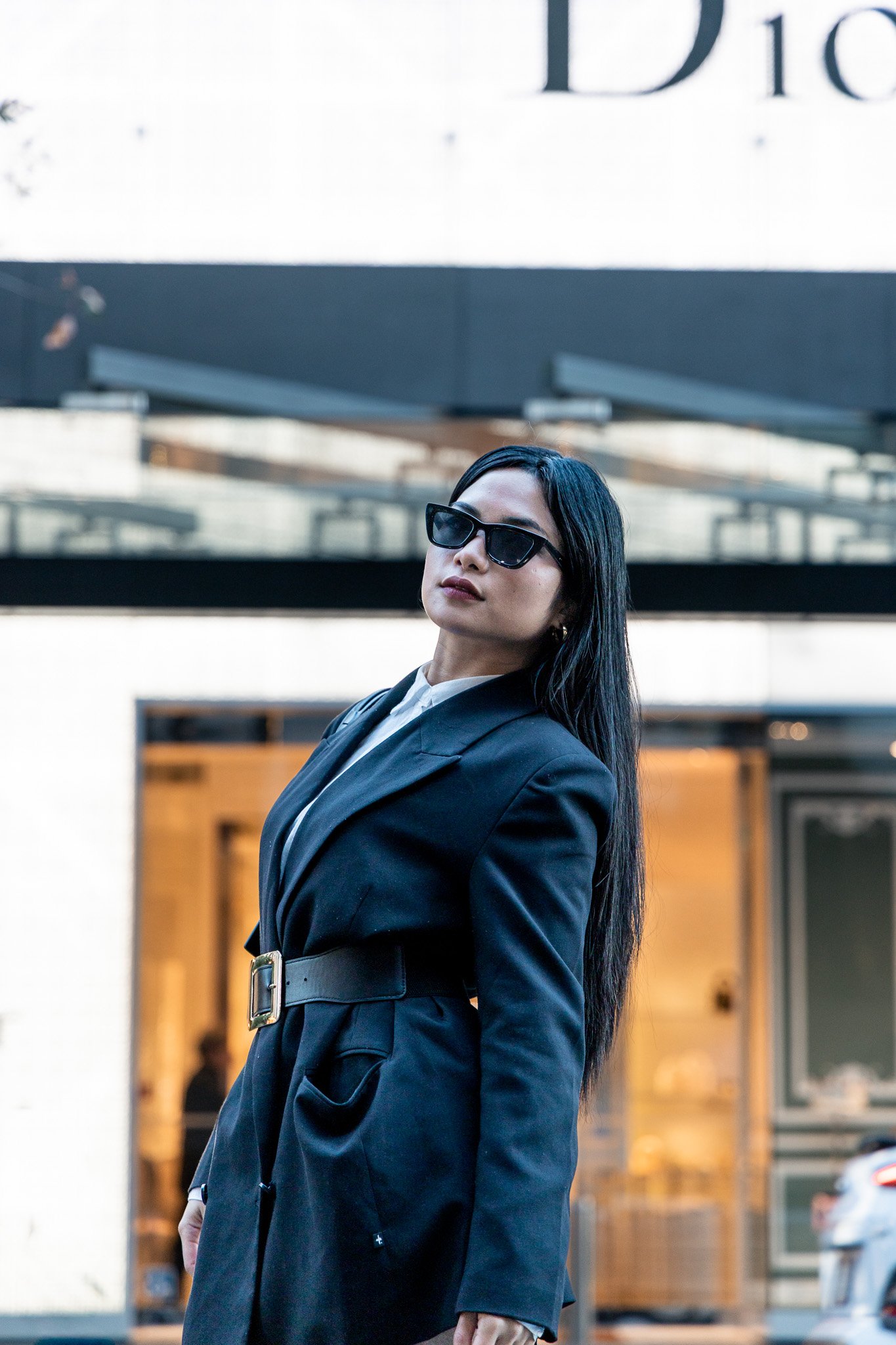 A woman with long black hair, wearing sunglasses, a black blazer, and a white shirt, stands outdoors in front of a modern building with large windows and a sign with the word "Dior" in the background.