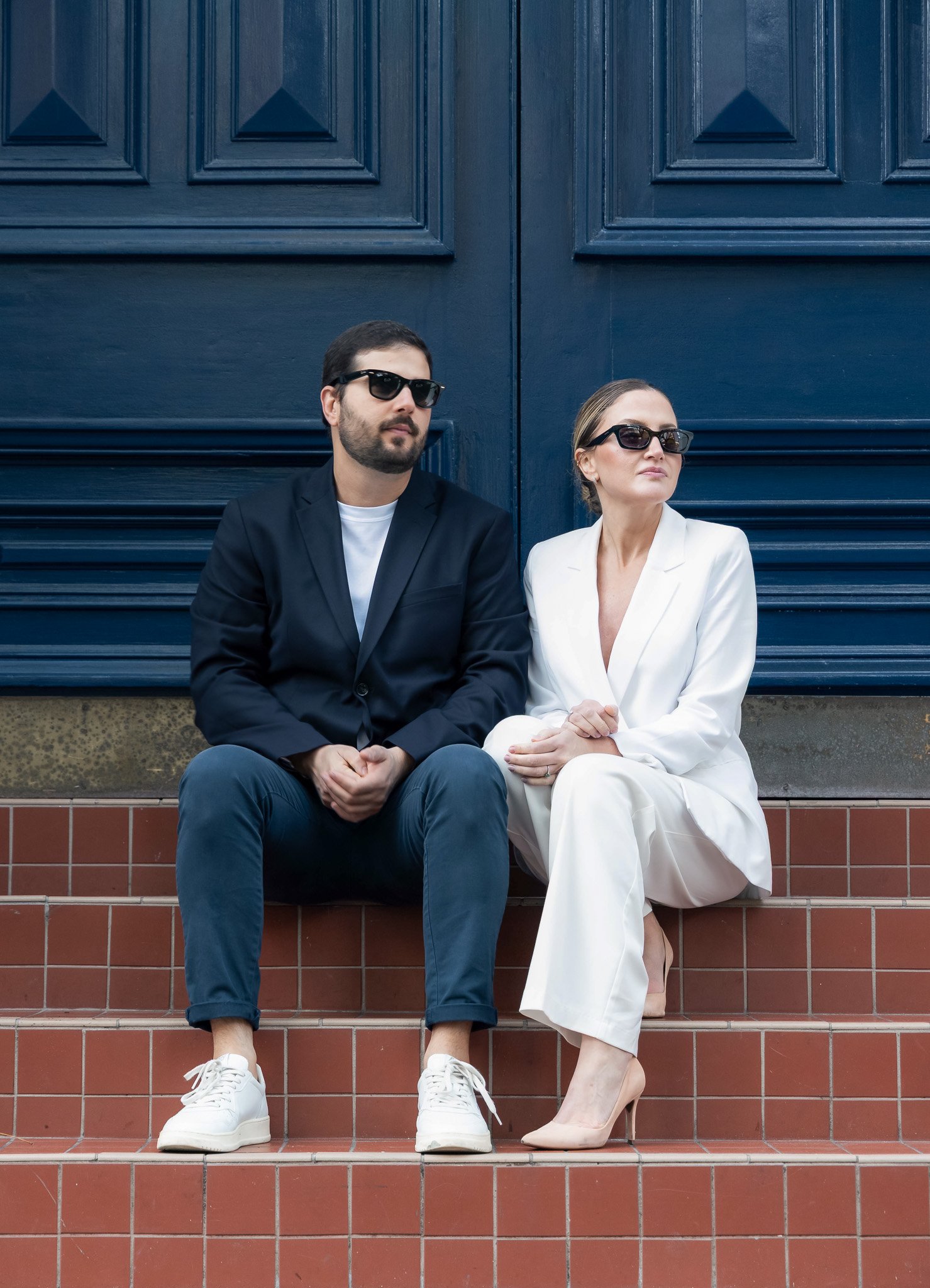 A man and a woman sitting on steps in front of a blue wall, both wearing black sunglasses. The man is dressed in a navy blazer and white sneakers, with a beard and dark hair. The woman is dressed in a white suit, beige heels, and has her hair styled 