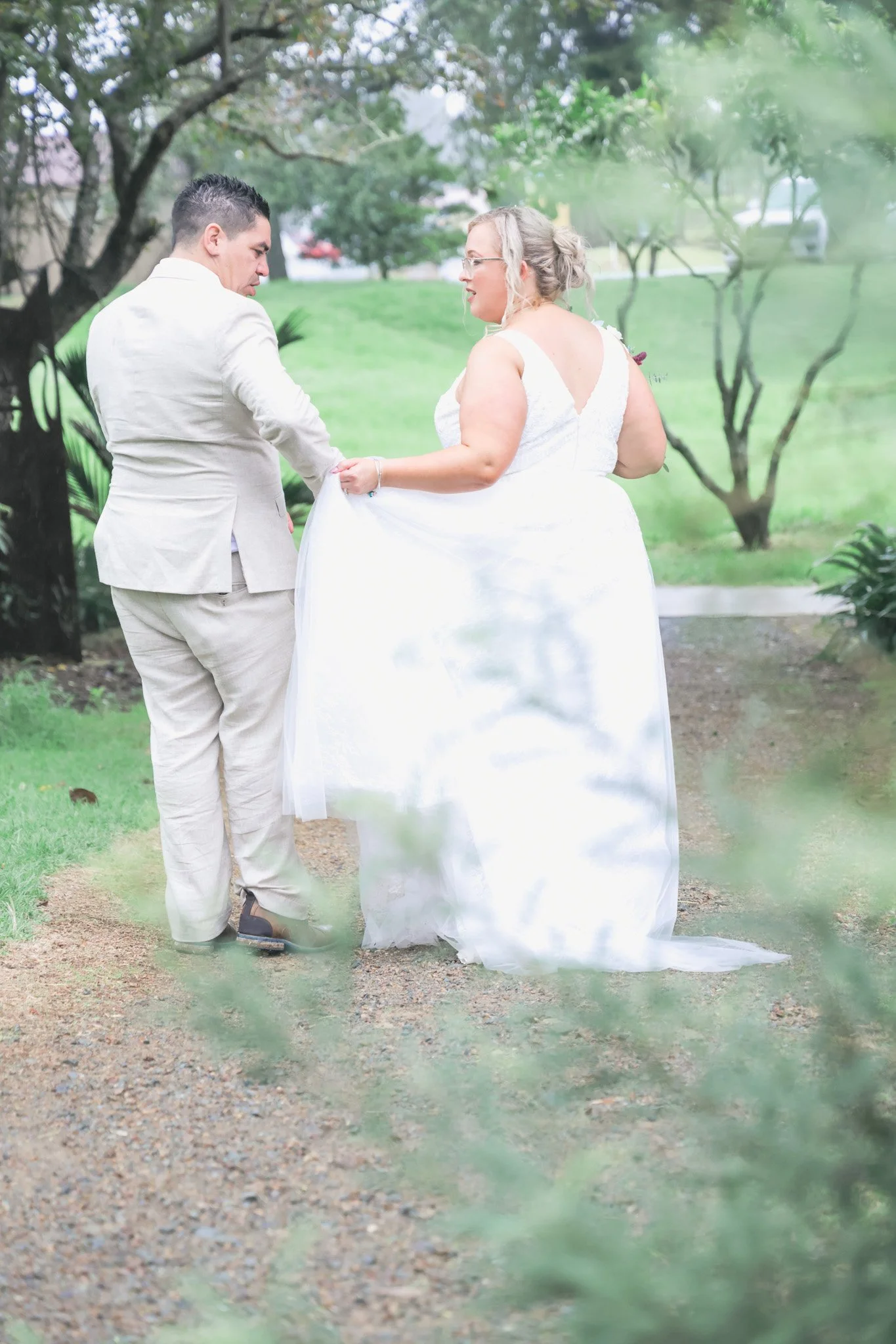 A couple dressed in wedding attire walking outdoors in a park-like setting with trees and green grass.