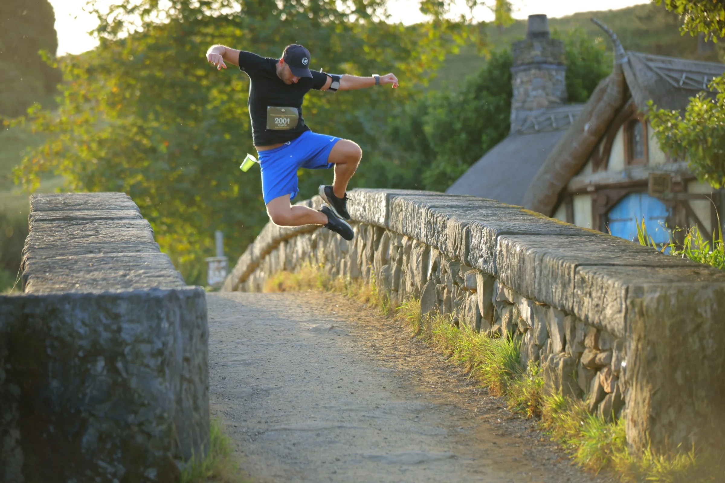 A man jumping over a stone wall on a dirt path in a rural area during sunset, with green trees and a thatched cottage in the background.