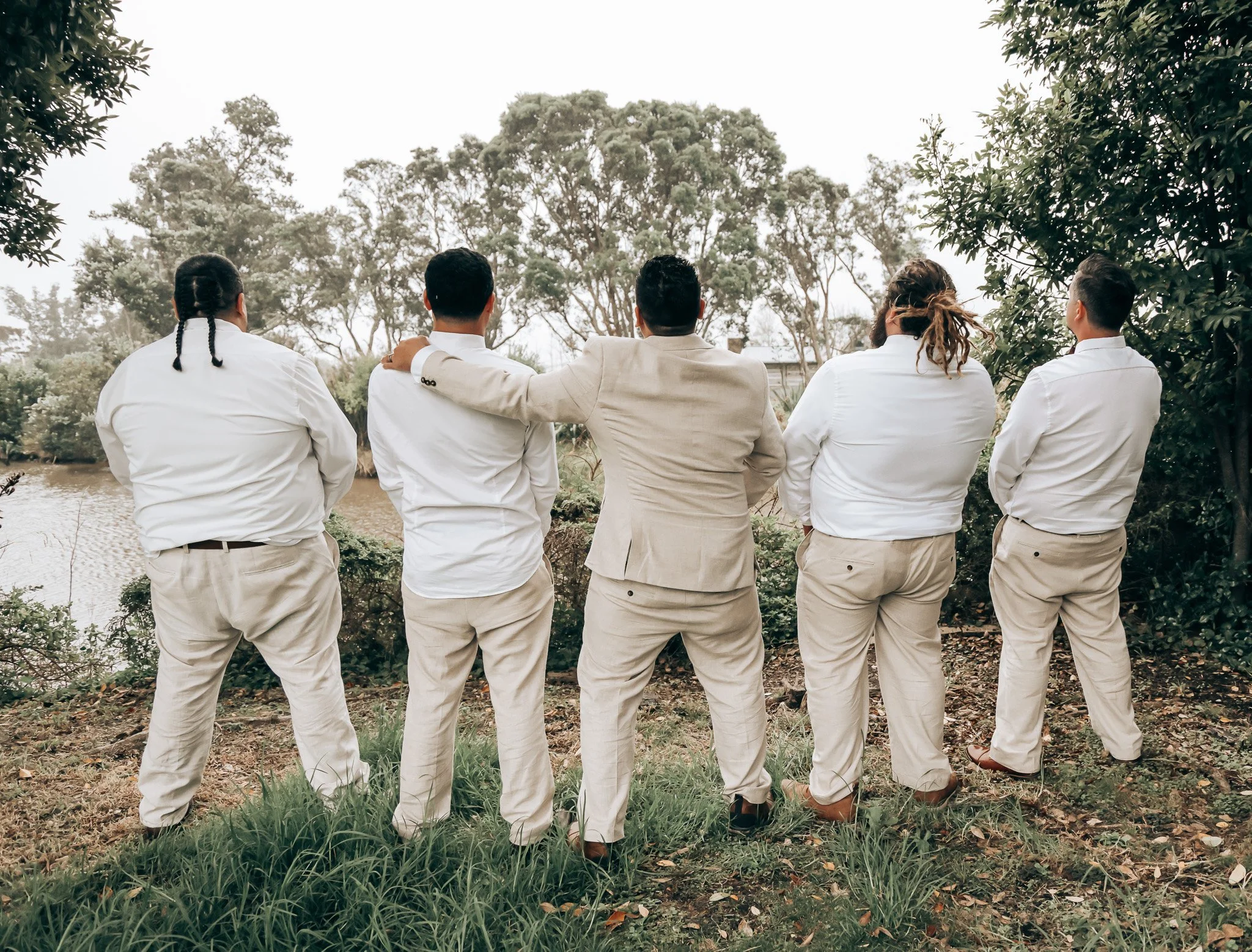 Six people in formal attire stand outdoors near water, facing away from the camera, with arms around each other's shoulders, surrounded by trees.