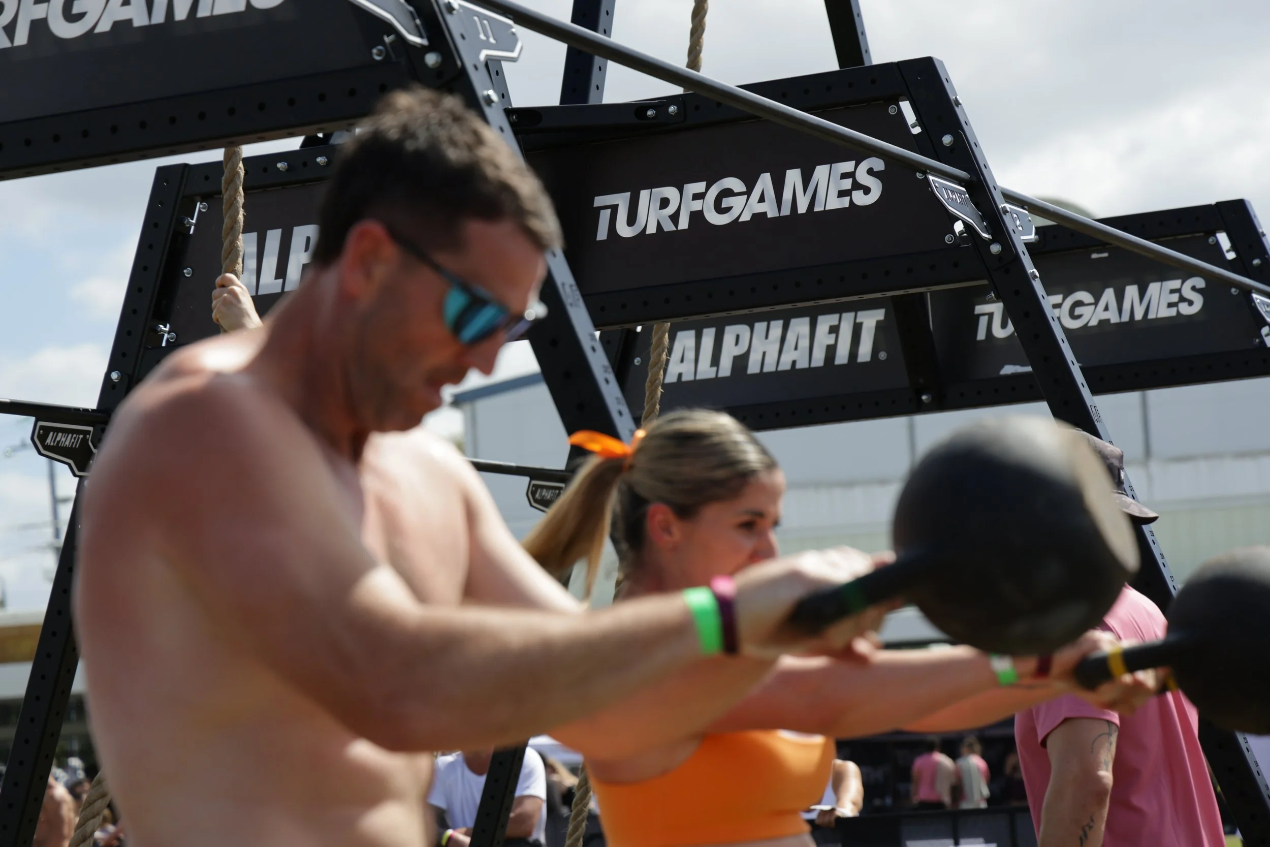People participating in an outdoor fitness event, with a focus on a shirtless man and others stretching or exercising, under black structures labeled 'TURFGAMES' and 'ALPHAFIT'.