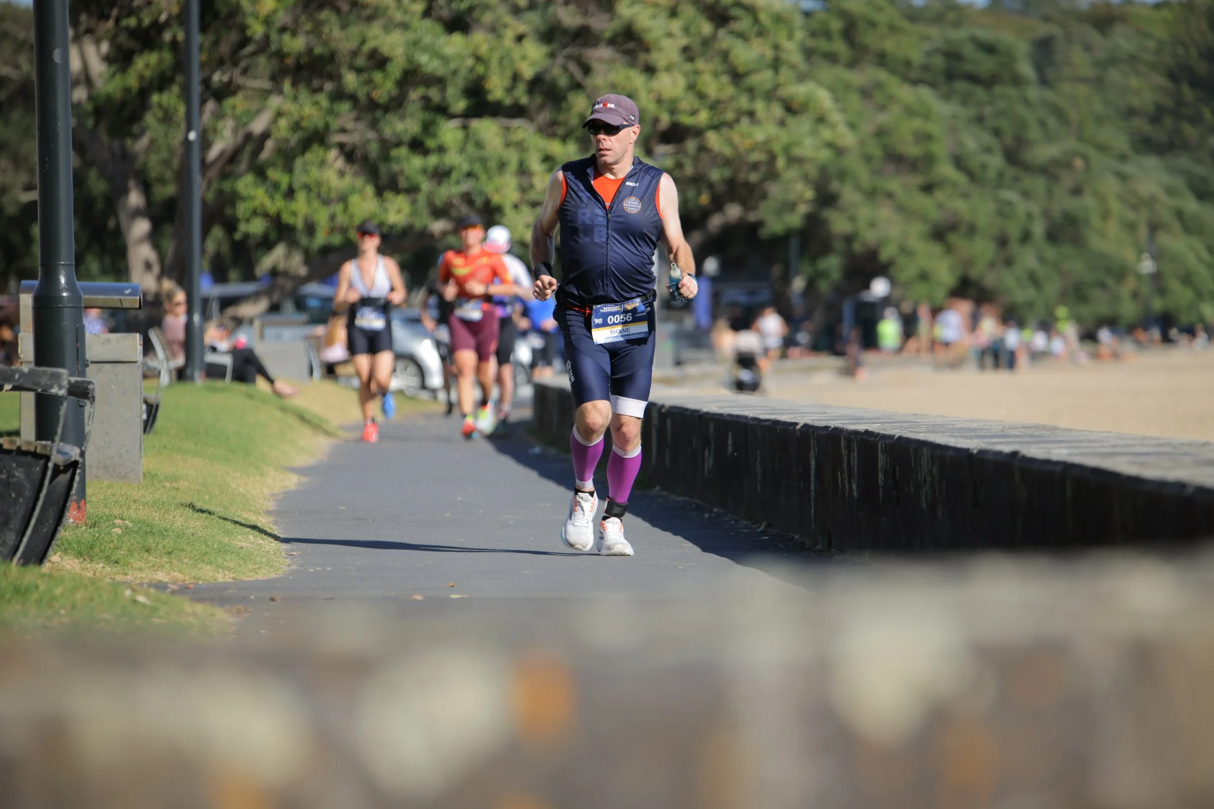 A man running in a race along a paved path with other runners behind him in a park with trees.