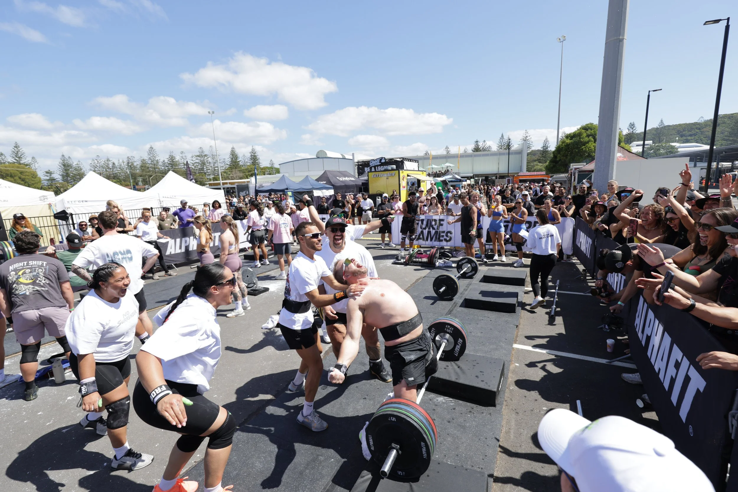 A large outdoor event with many people gathered around a weightlifting competition. Two men are engaged in a lift, with one assisting the other. The crowd appears excited, cheering and taking photos. Tents and banners are visible in the background on