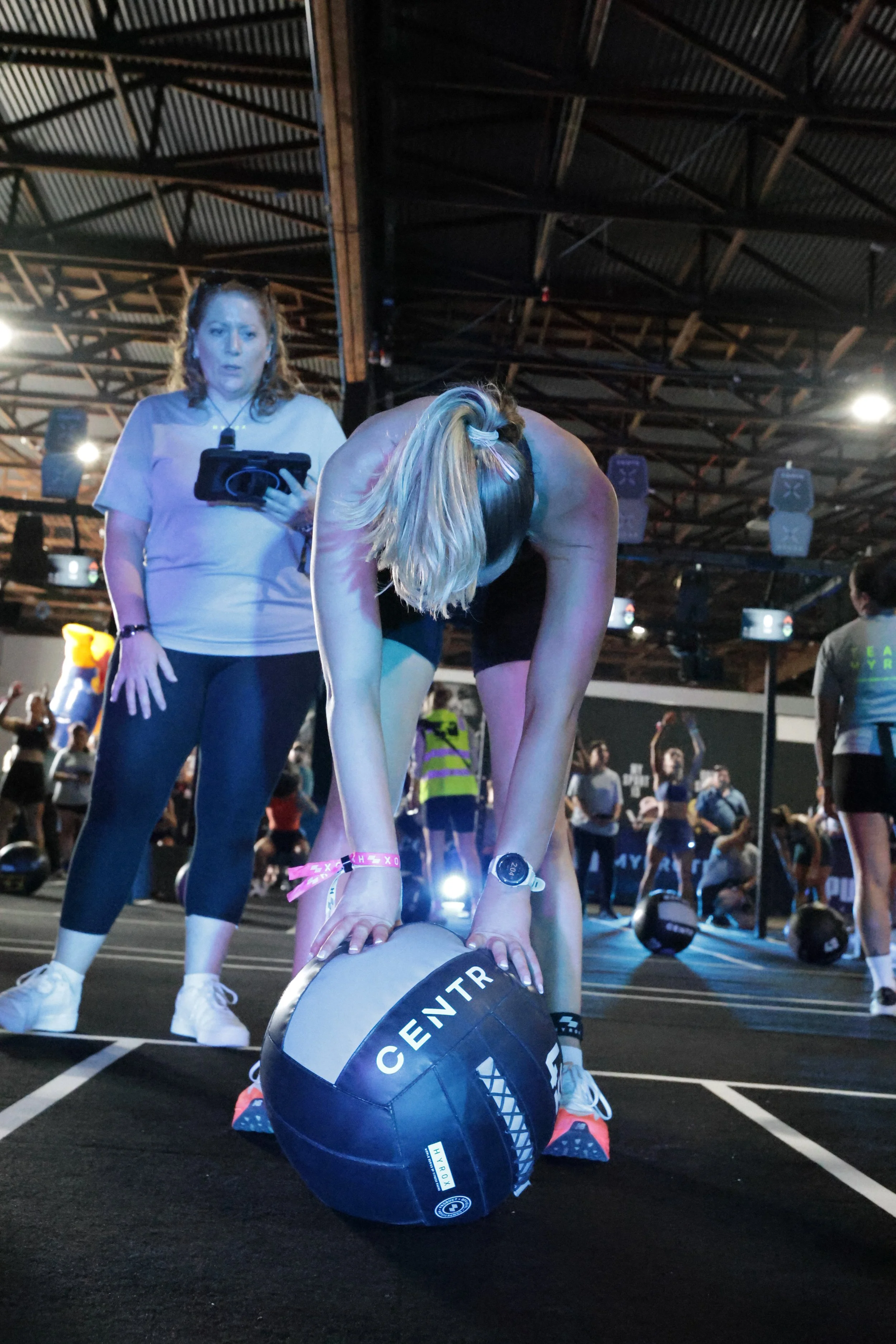 A woman is performing a plank exercise on a gym floor while another woman watches and holds a tablet. The scene is inside a gym or fitness event with spectators and equipment visible.