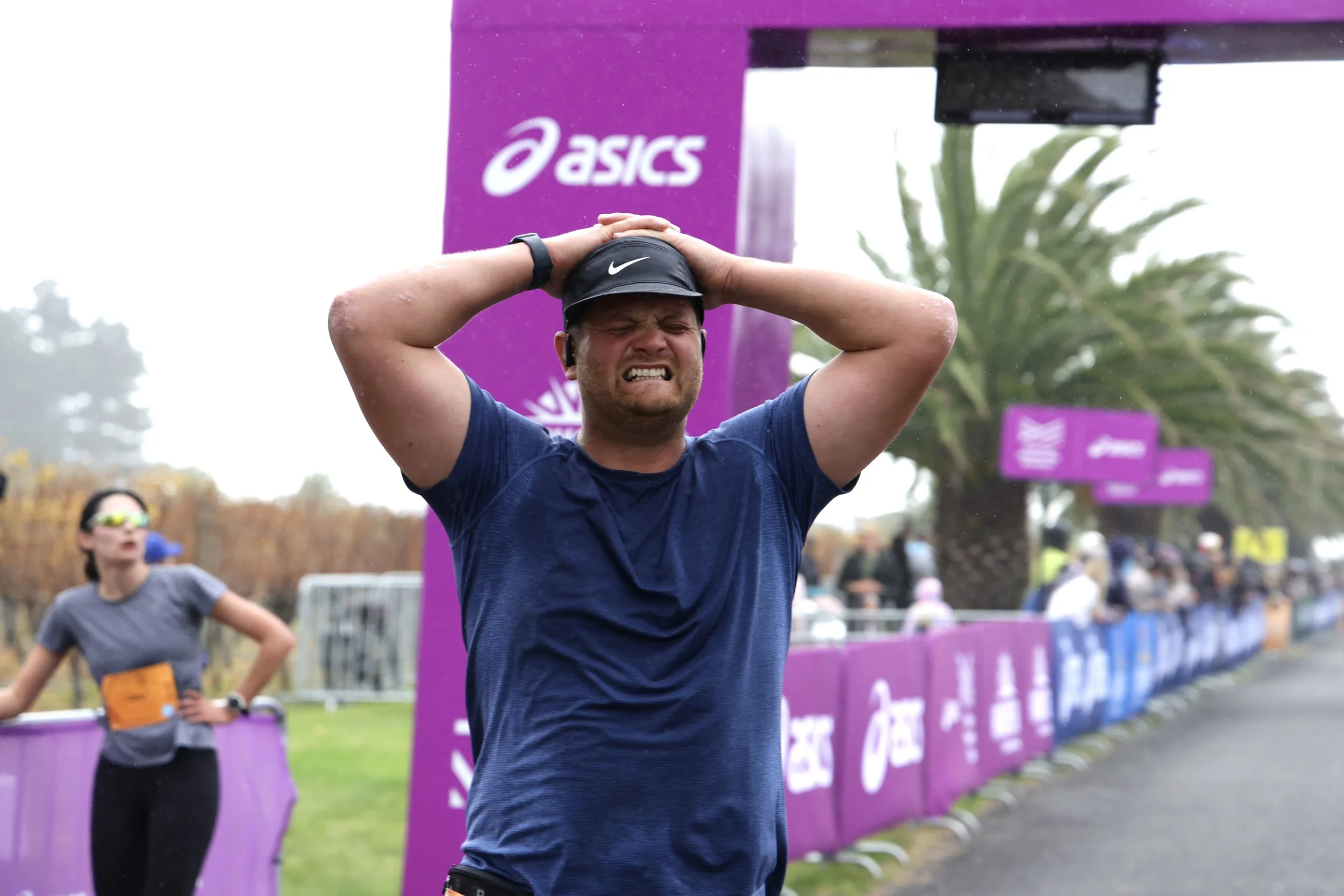 A man wearing a black Nike hat and a blue shirt holds his head with a pained expression during a running event. Behind him, a woman in sunglasses and a gray shirt is standing with her hand on her hip. The background features purple banners with white