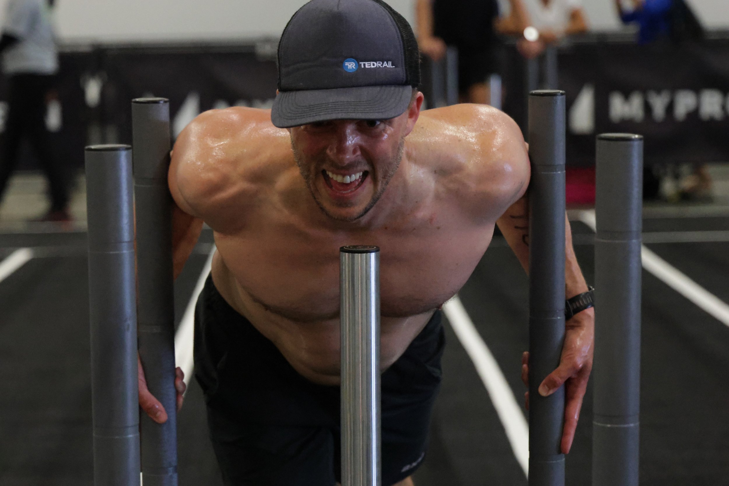 A shirtless man wearing a black cap with an embroidered logo, pushing a sled with metal bars in a gym.