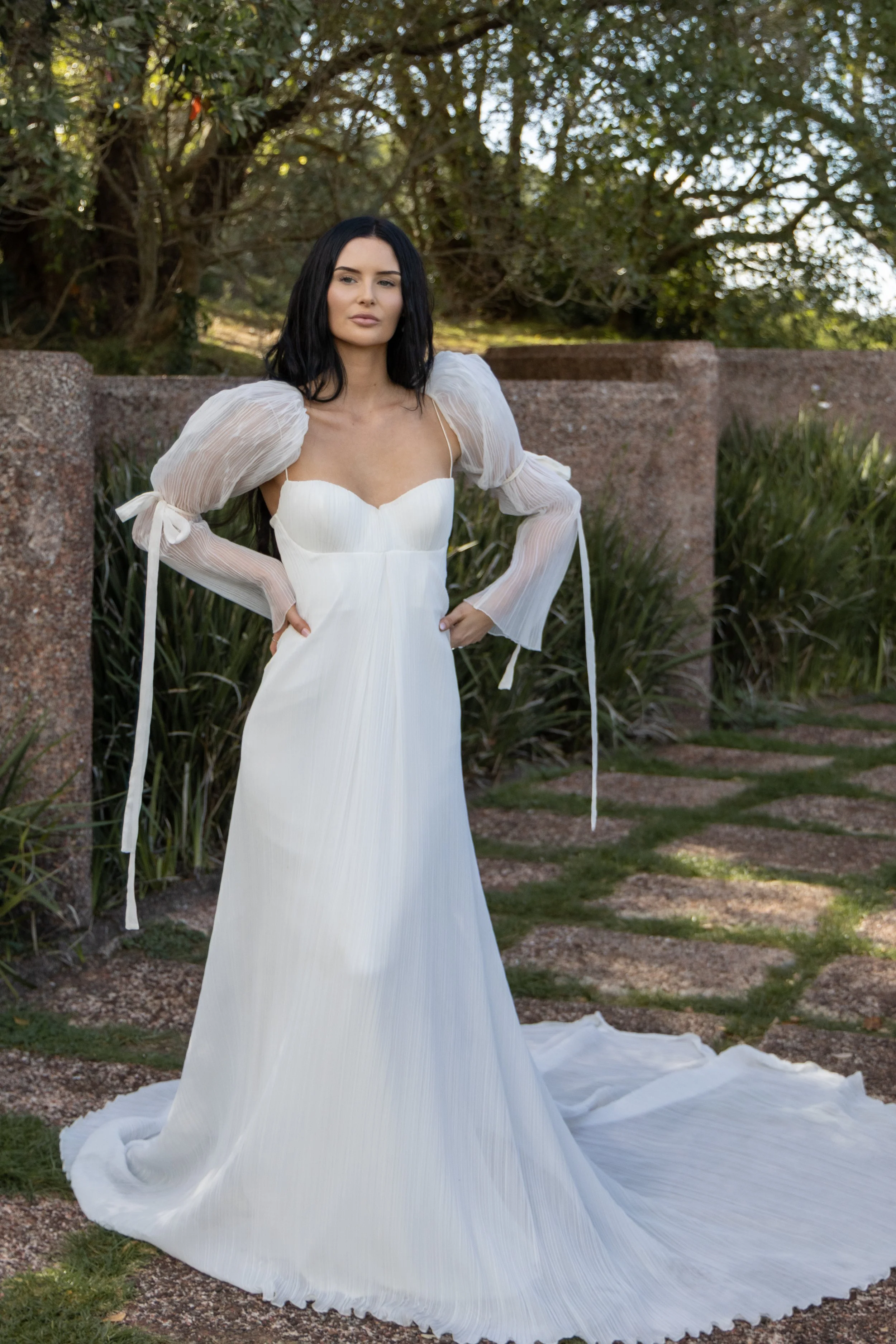 Woman in a white wedding dress with puffed sleeves standing outdoors near a stone wall and green foliage.
