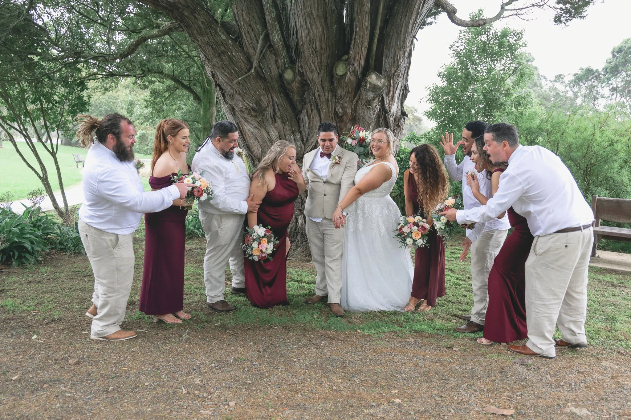 Wedding party standing outdoors under a large tree, with the bride and groom at the center, surrounded by bridesmaids and groomsmen smiling and celebrating.